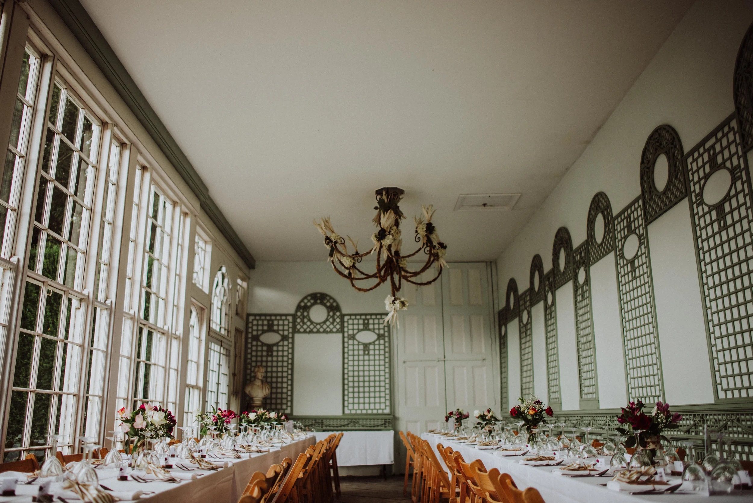 Long dining table set with white tablecloth, floral centerpieces, and glassware in an elegant room with large windows and decorative wall panels, chandelier hanging from the ceiling.
