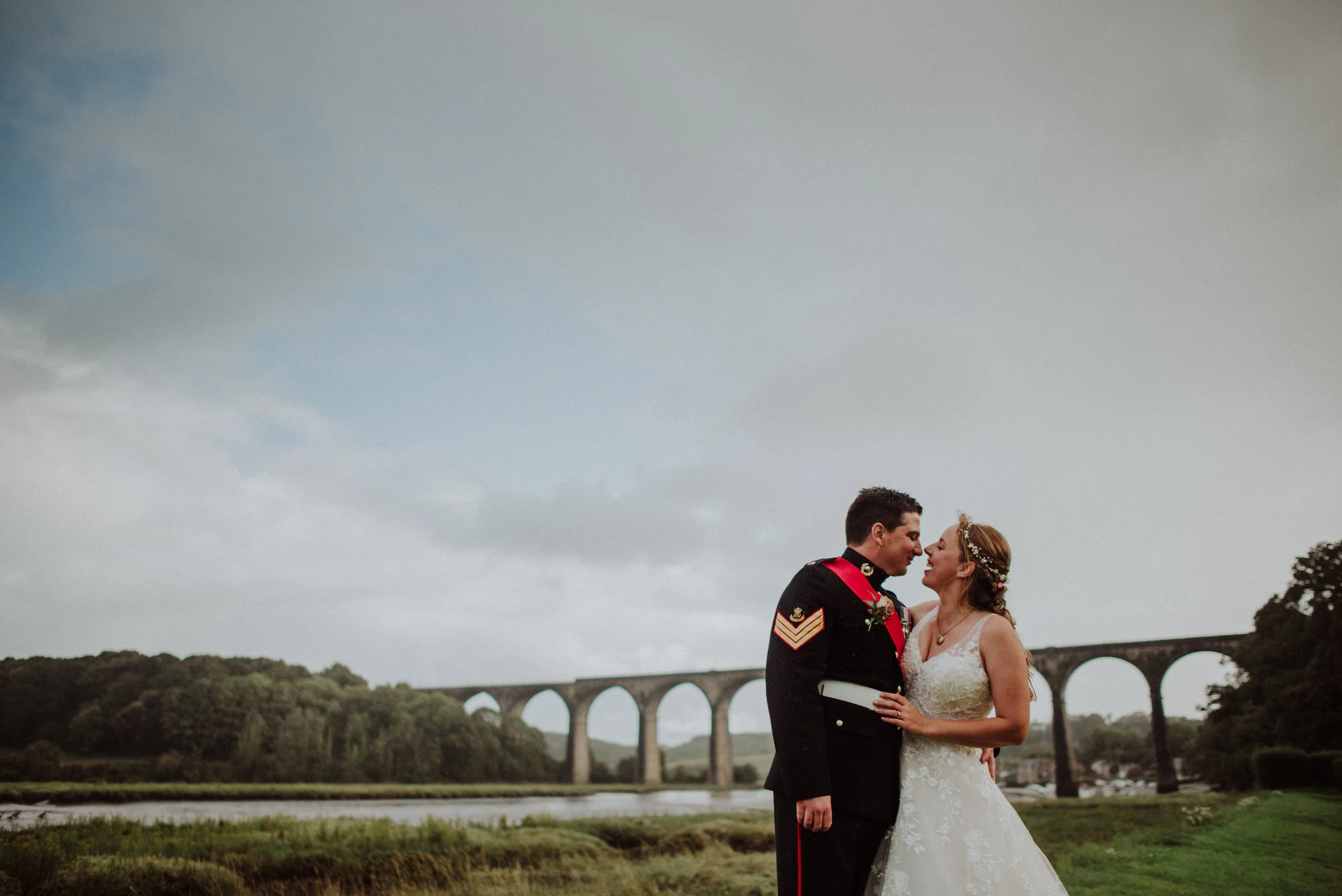 A bride and groom share a kiss outdoors near a river with a large stone bridge in the background.