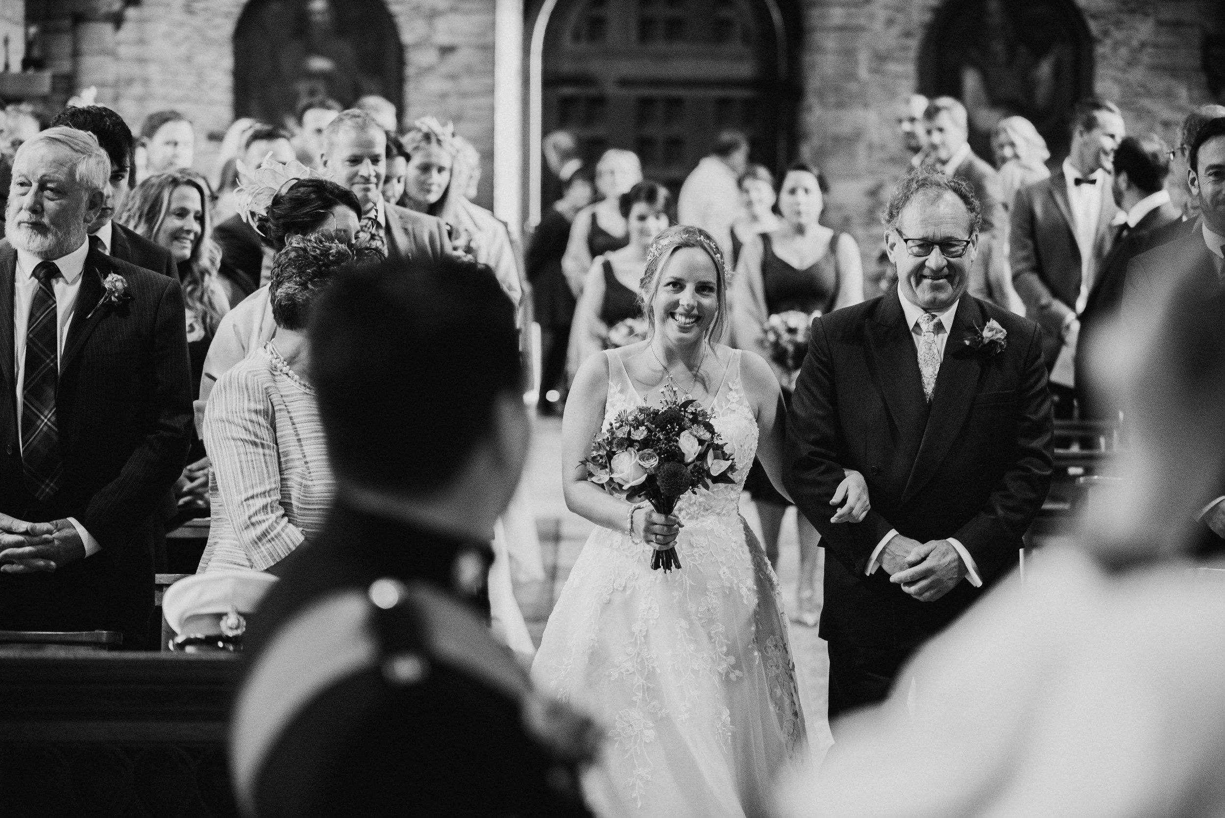 A bride walking down the aisle in a church, holding a bouquet, smiling as she is escorted by a man in a suit. Guests are seated and standing in the background, watching and smiling.