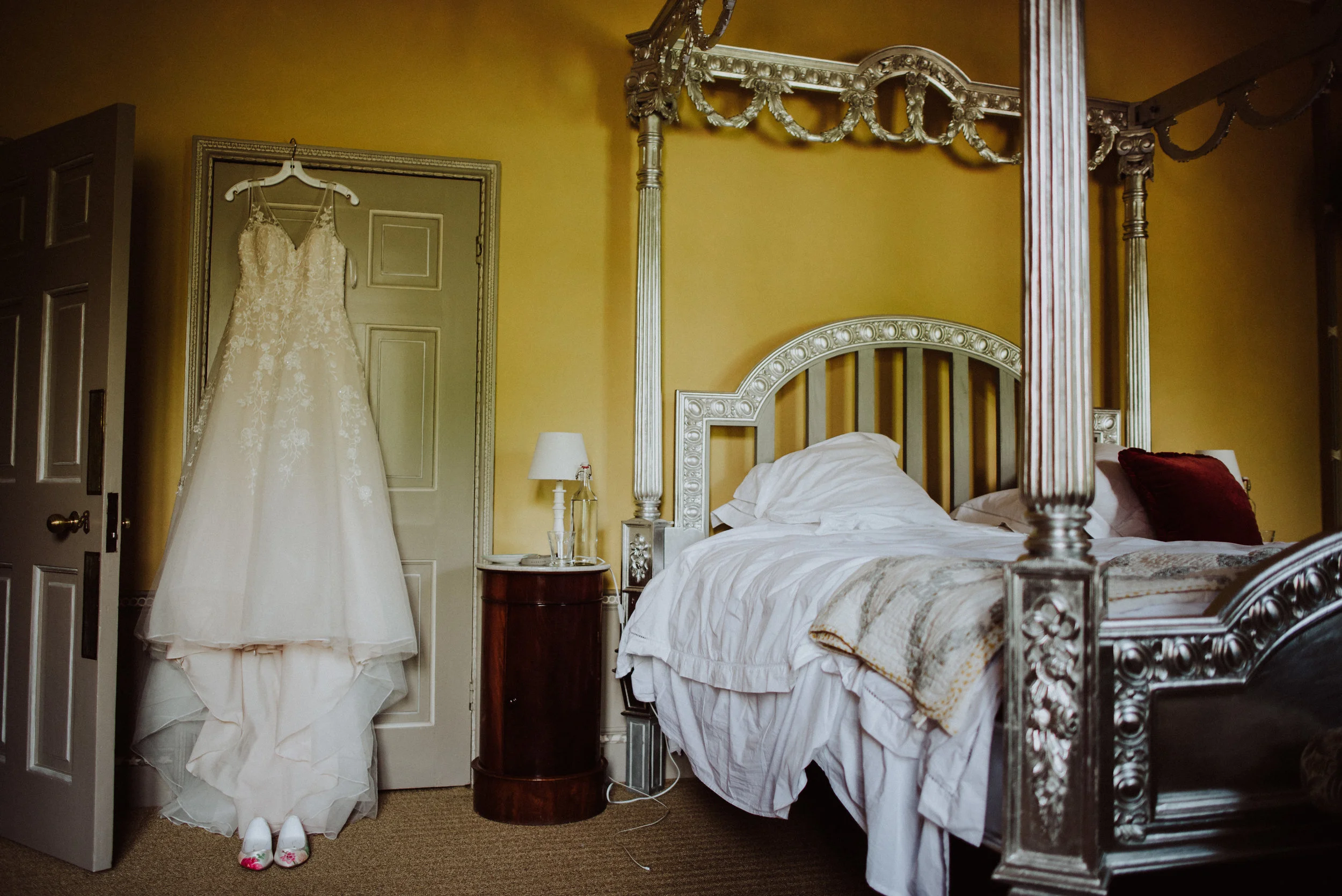 Wedding dress hanging on a door in a bedroom with a large ornate bed and nightstand.