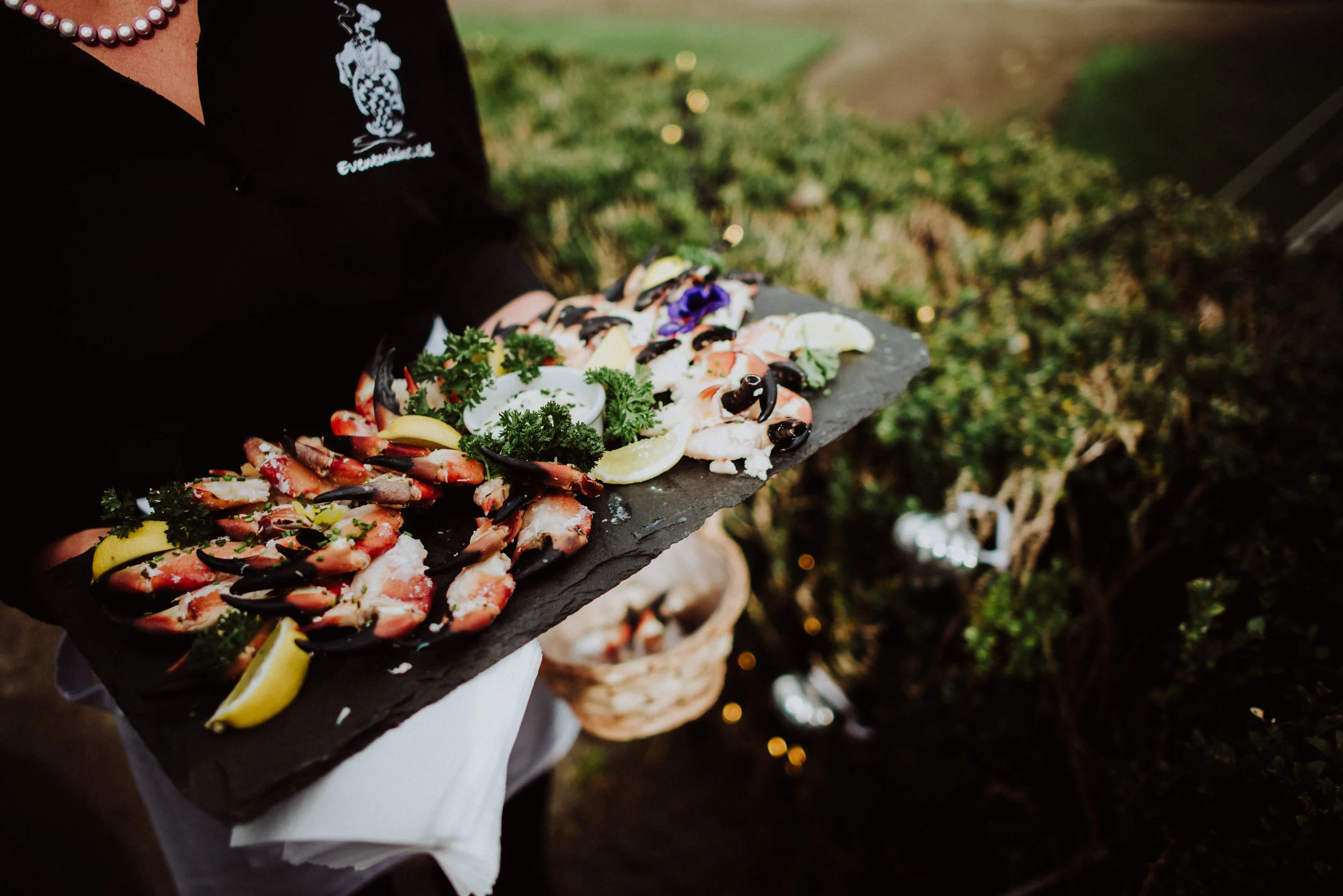 A person holding a black slate platter with an assortment of seafood, including shrimp and crab legs, garnished with lemon wedges, parsley, and a dipping sauce, outdoors near greenery.