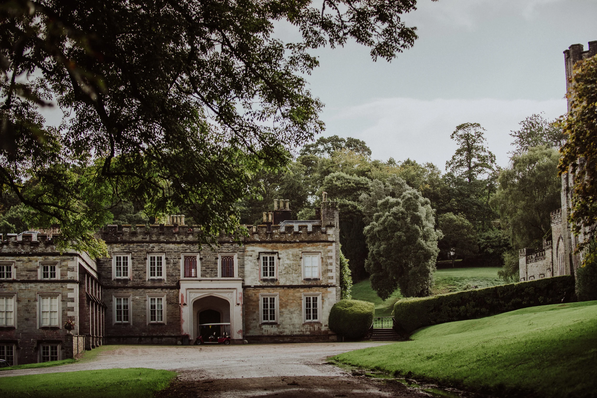 A historic stone castle or mansion with a gravel driveway, surrounded by lush green manicured lawns and tall trees, under an overcast sky.
