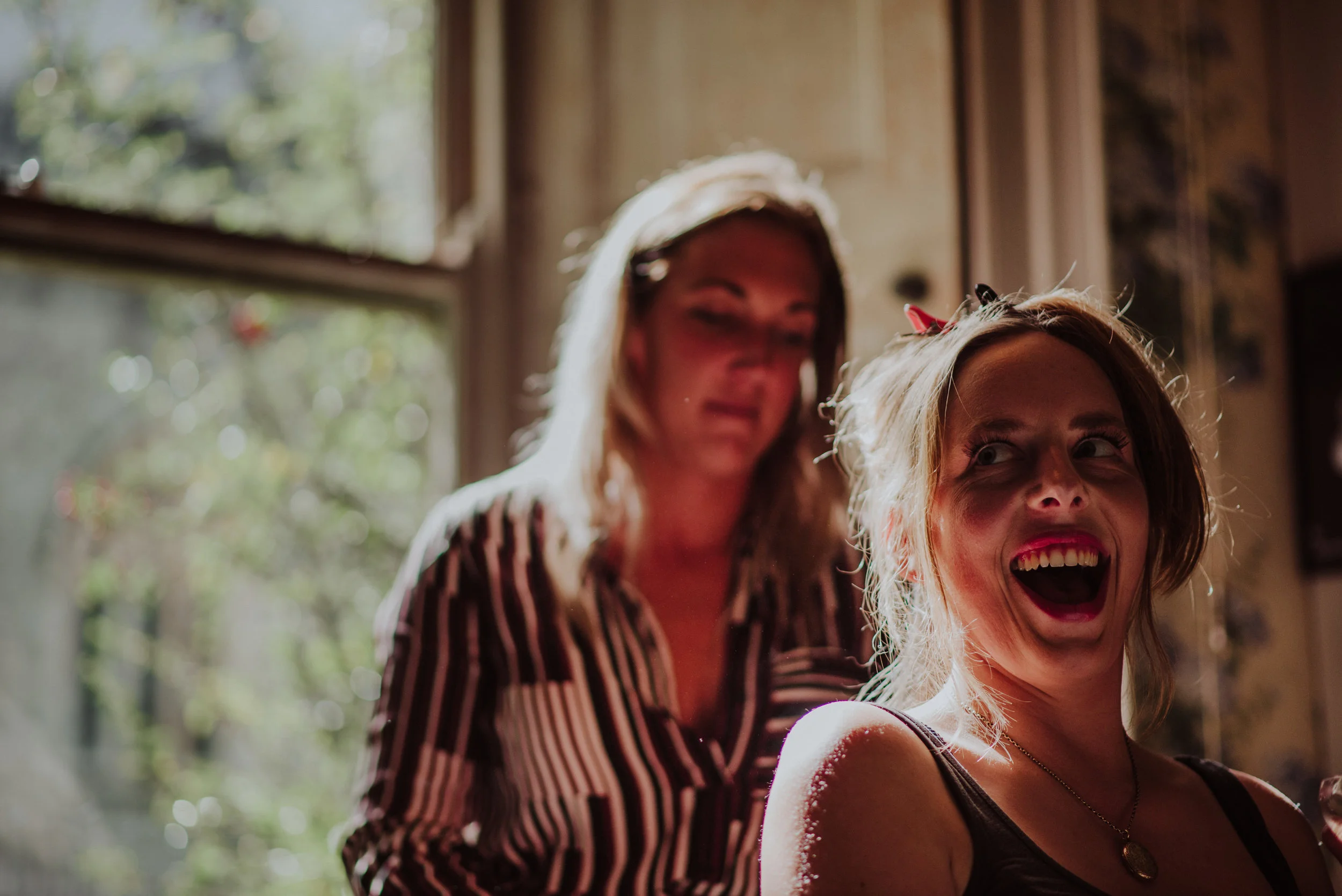 Two women with light skin are inside a room near a window with sunlight coming in. One woman is smiling and appears to be having fun; she has shoulder-length hair, is wearing a black top and a necklace. The other woman is slightly blurred, standing b
