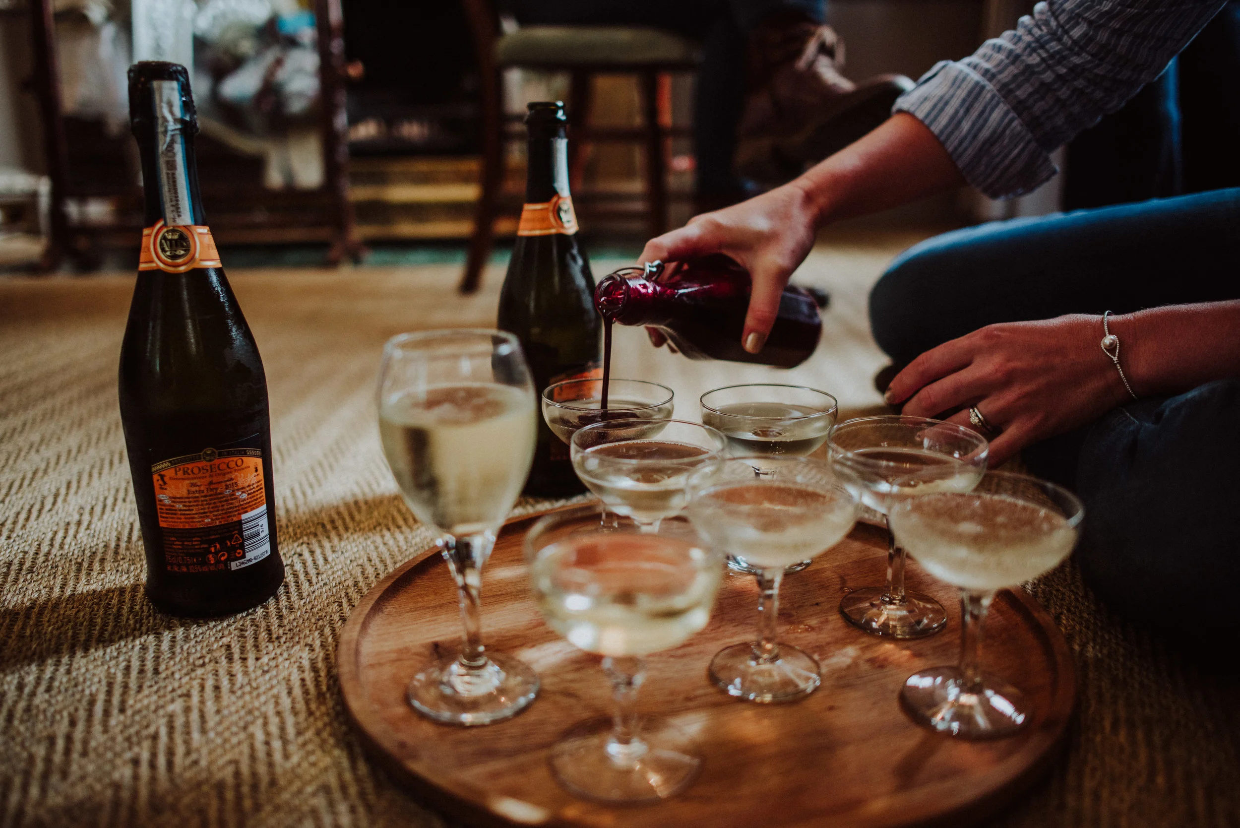 Person pouring red wine into glasses on a wooden tray with bottles of Prosecco nearby, in a cozy indoor setting.