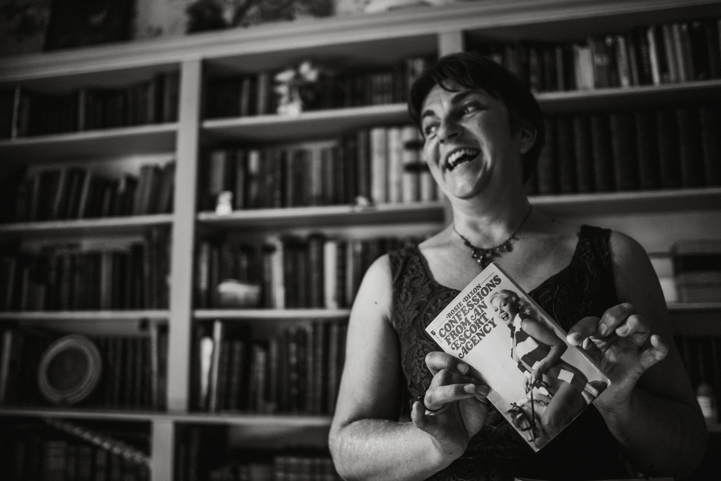 A woman laughing and holding a book titled 'Confessions from an Escort Agency' in front of a background filled with bookshelves.