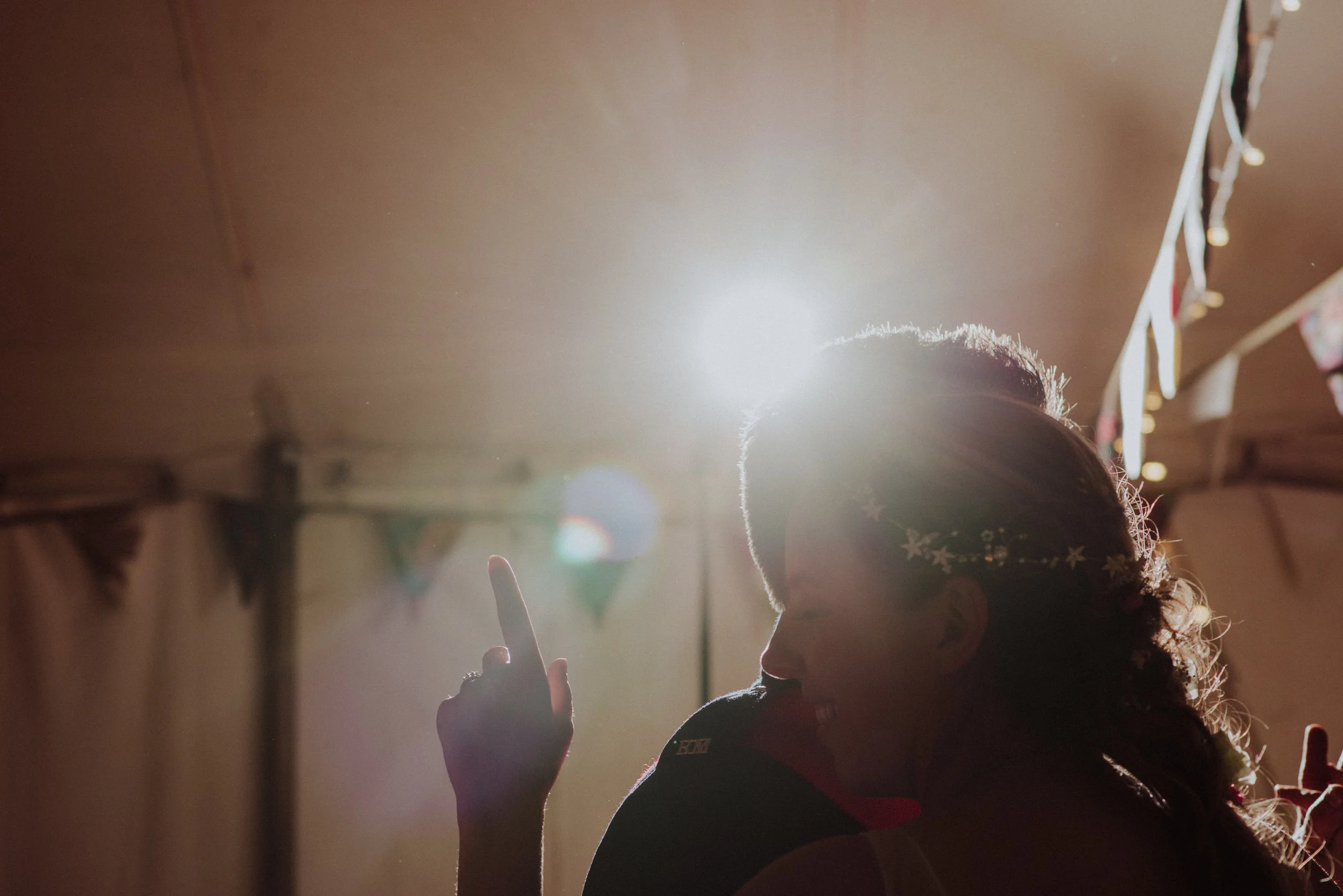 A silhouette of a person with a flower headband, smiling and pointing up, backlit by bright sunlight inside a decorated tent.