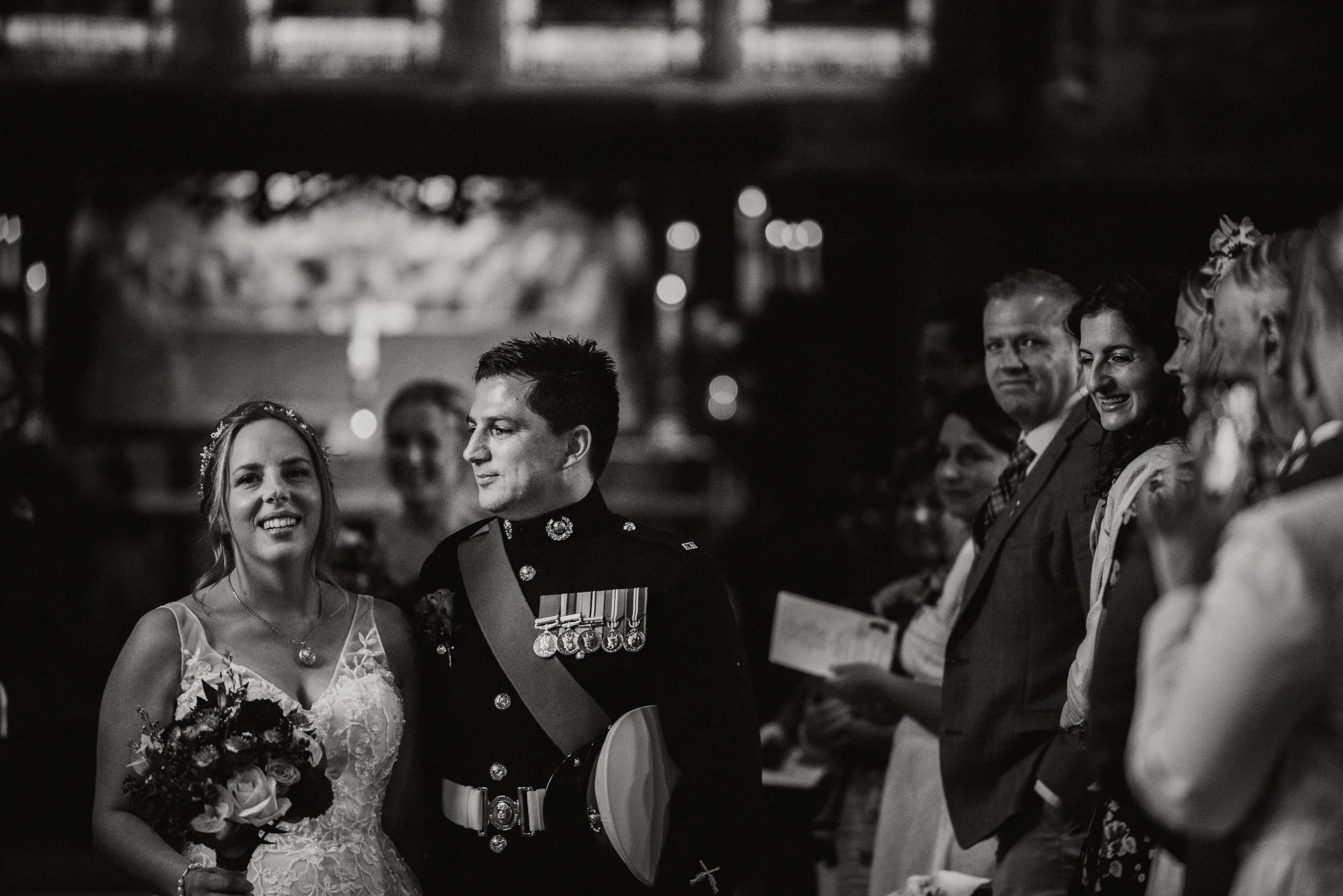 Black and white photo of a wedding ceremony with a bride and groom, surrounded by guests, indoors at night.