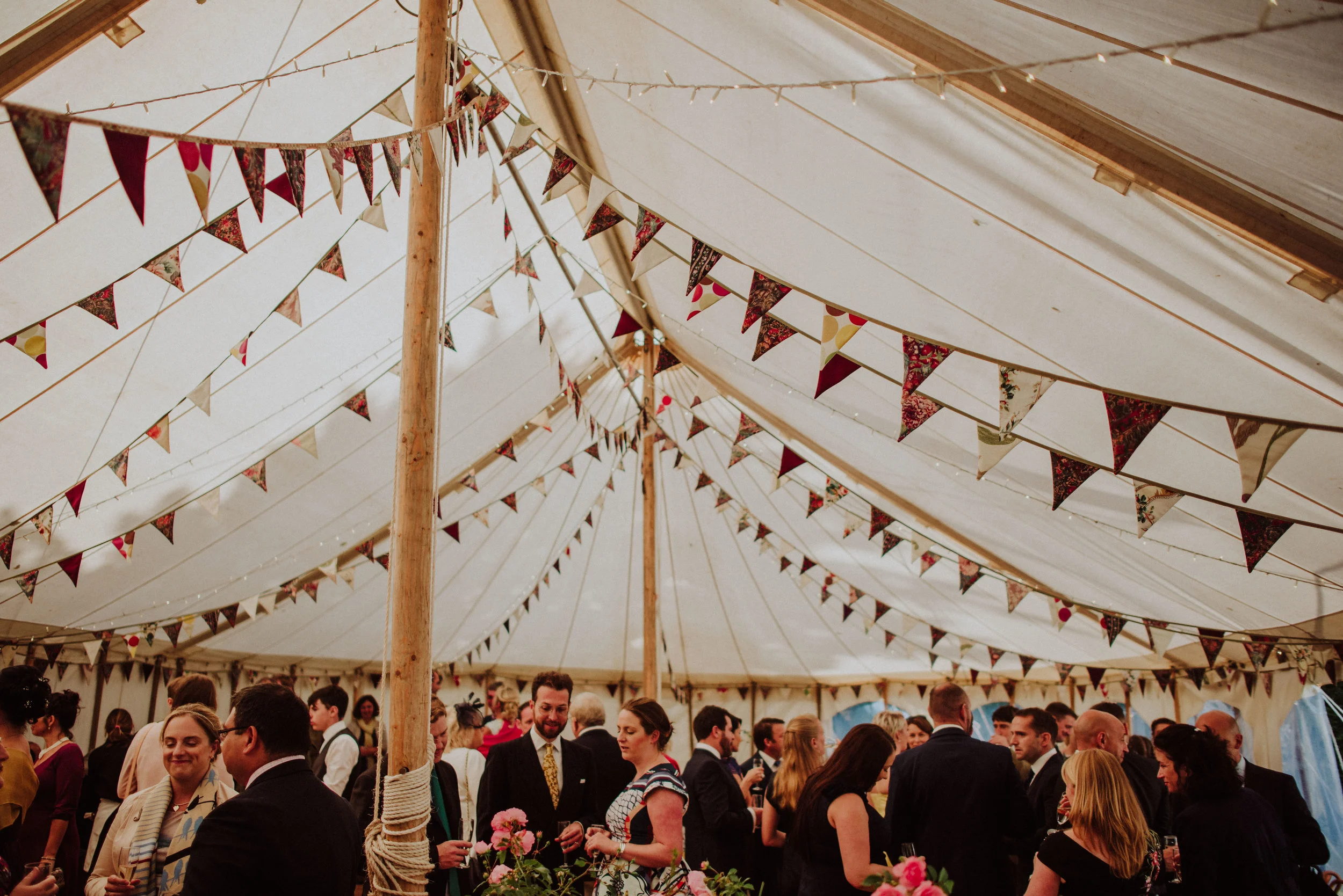 People dressed in formal attire socializing inside a decorated event tent with bunting flags and floral arrangements.