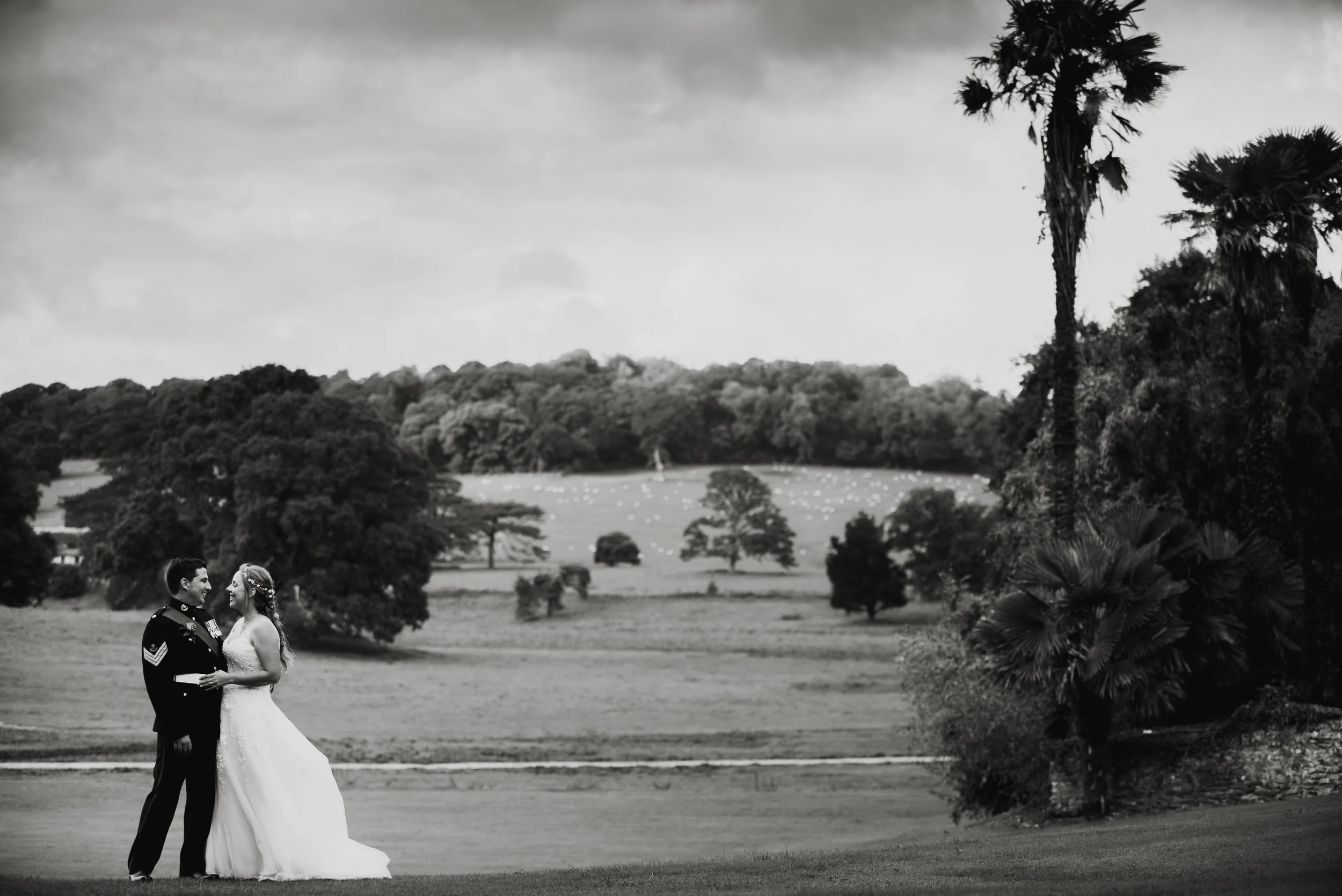 A black and white photograph of a bride and groom standing and gazing at each other in a park with trees and rolling hills in the background.
