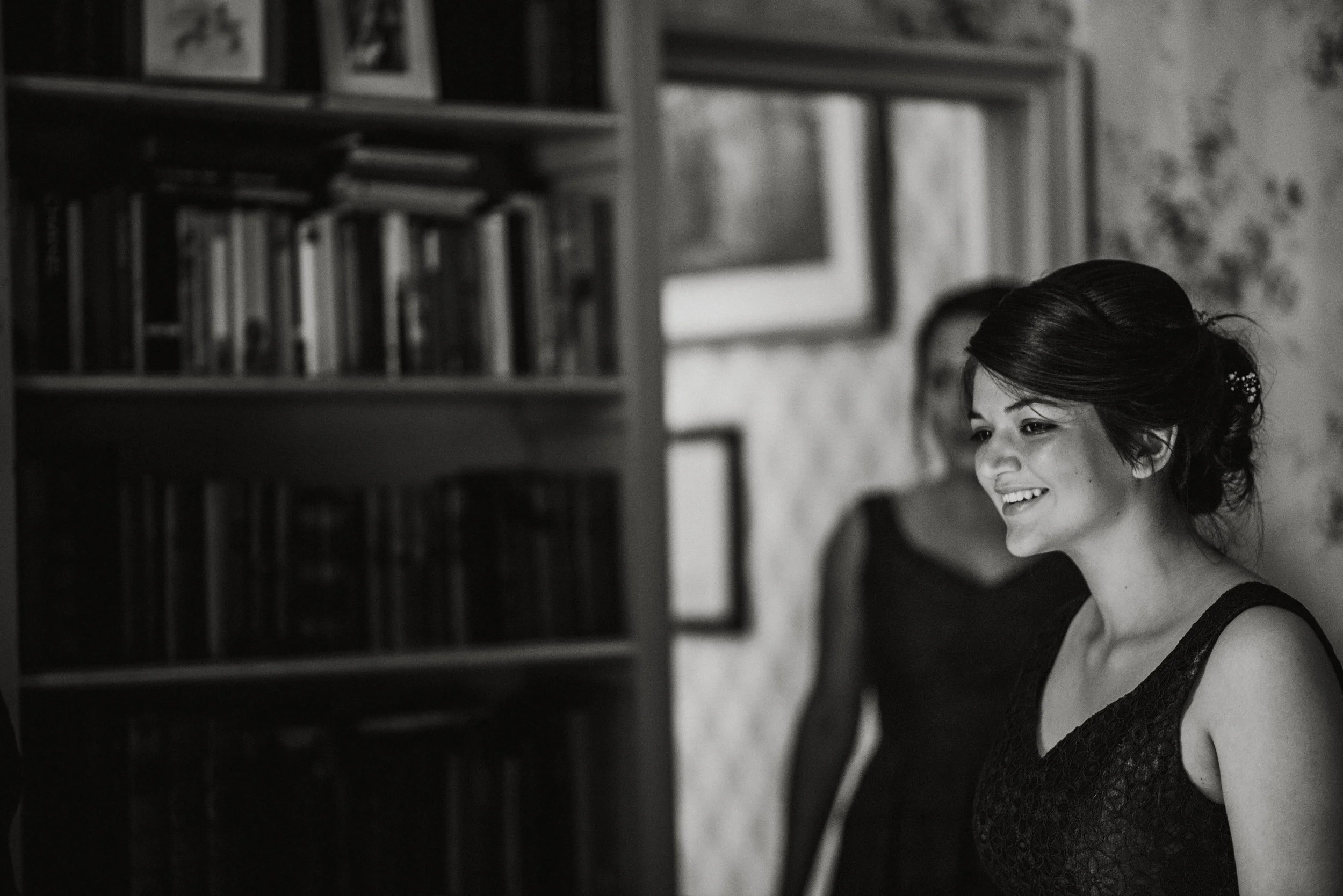 A young woman with dark hair styled in an updo, smiling, is in a room with a bookshelf and framed pictures on the wall. Another person, blurred, is in the background.