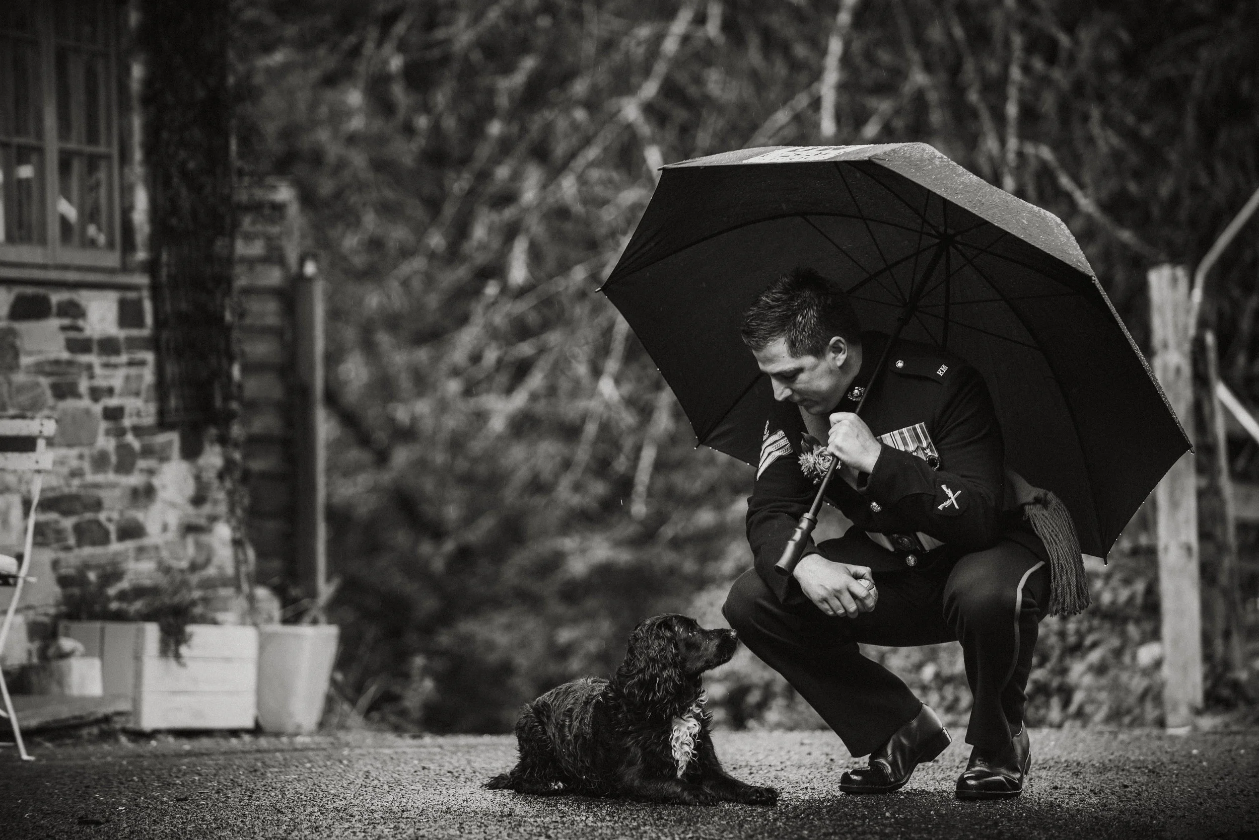 A man in a police uniform squatting with a umbrella, looking at a curled up dog on the ground outdoors on a rainy day.