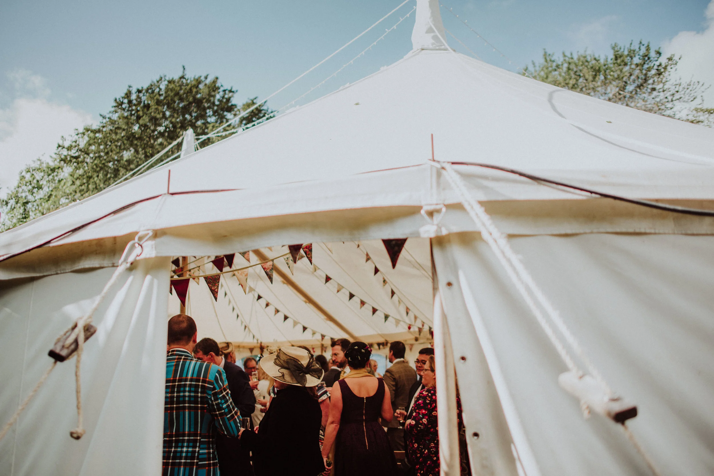 Crowd of people inside a decorated white event tent, with bunting flags hanging from the ceiling, outdoors on a sunny day with trees in the background.