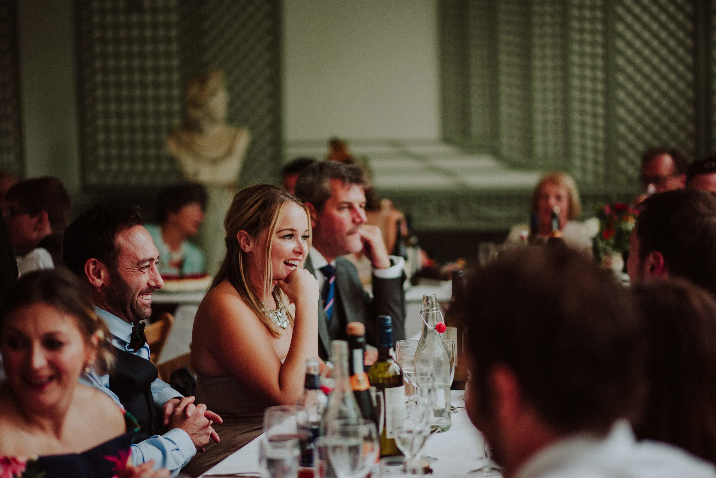 Guests at a formal event, sitting at a long table, laughing and enjoying the occasion, with bottles and glasses on the table, in an elegant room with classical art decor in the background.