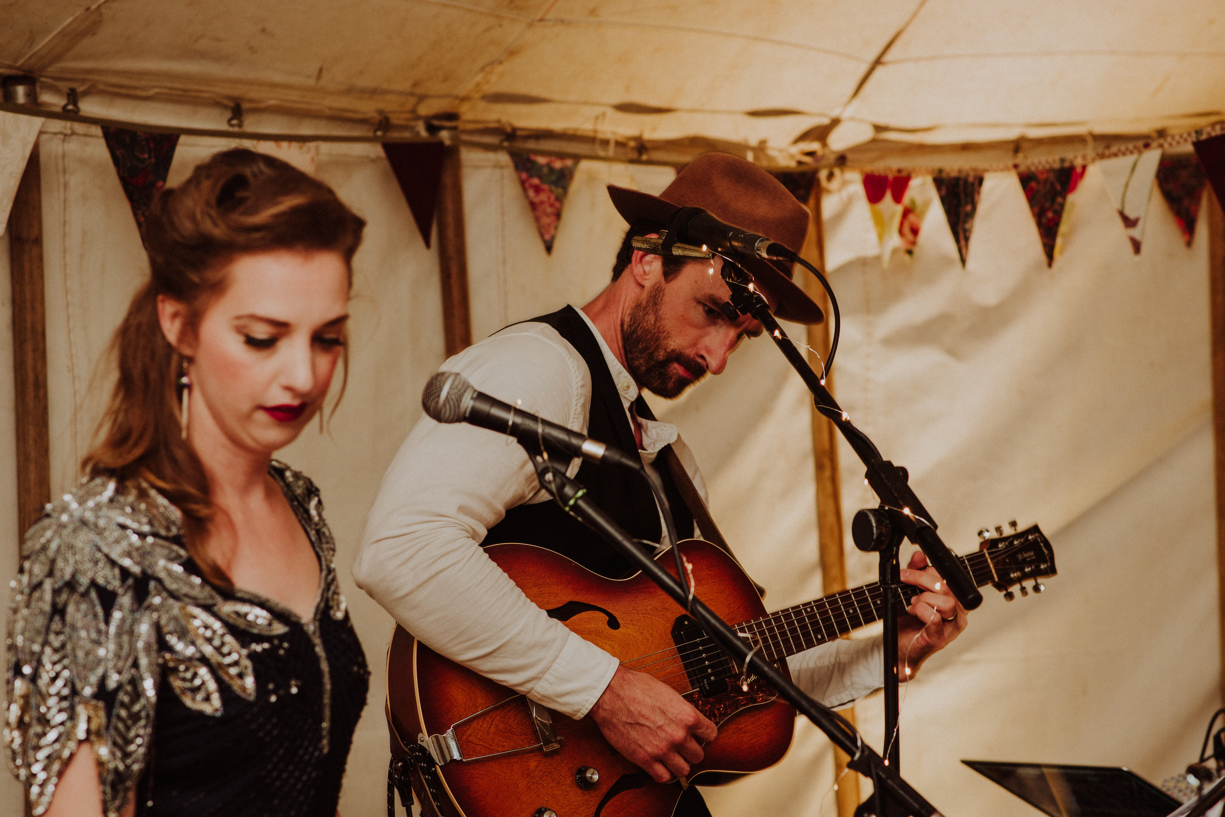 A woman and a man performing music indoors, with the woman looking down and the man playing an acoustic guitar while wearing a brown hat. Decorative bunting is hanging in the background.