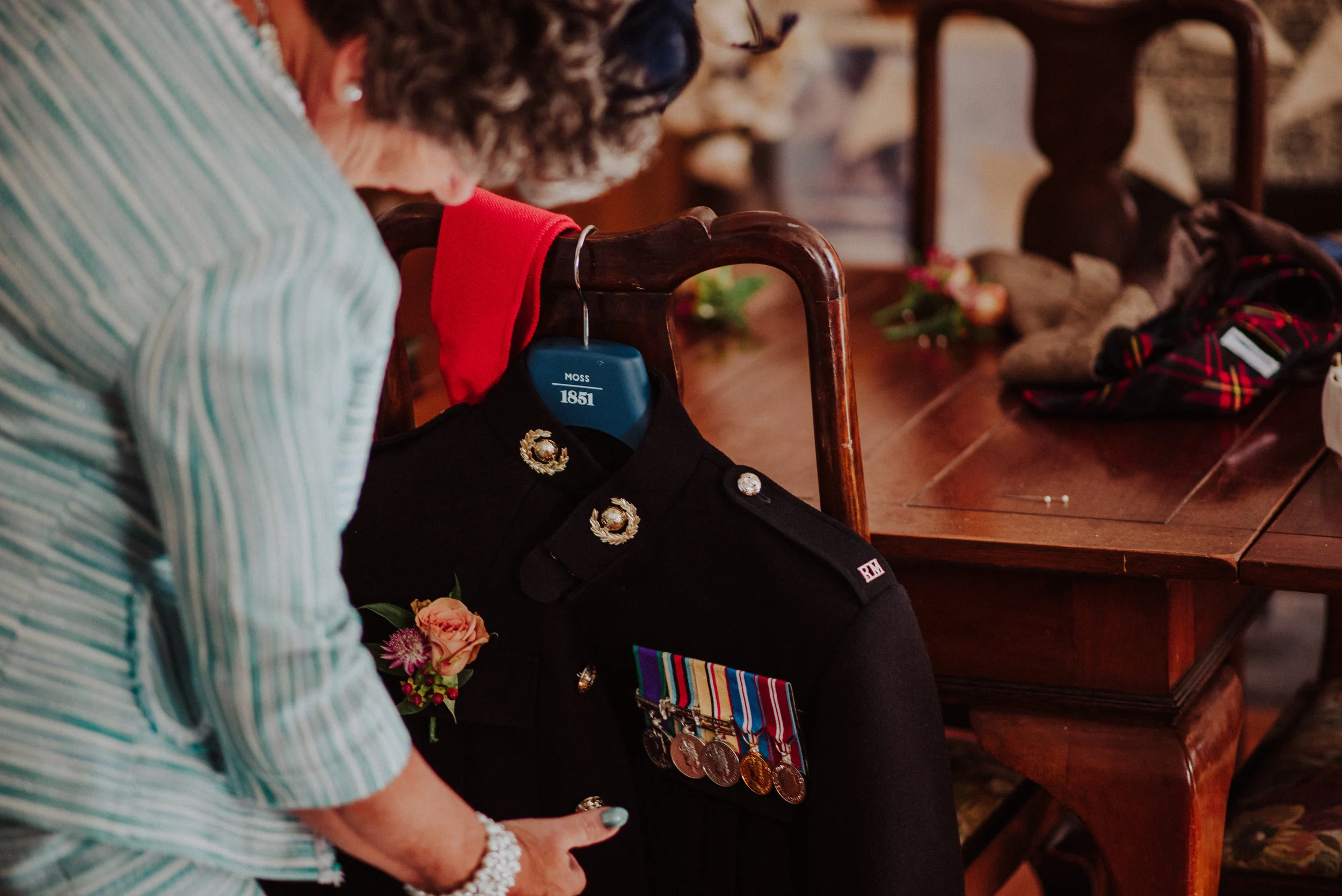 A woman in a light green striped shirt adjusting a black military uniform with medals and a pink flower boutonniere