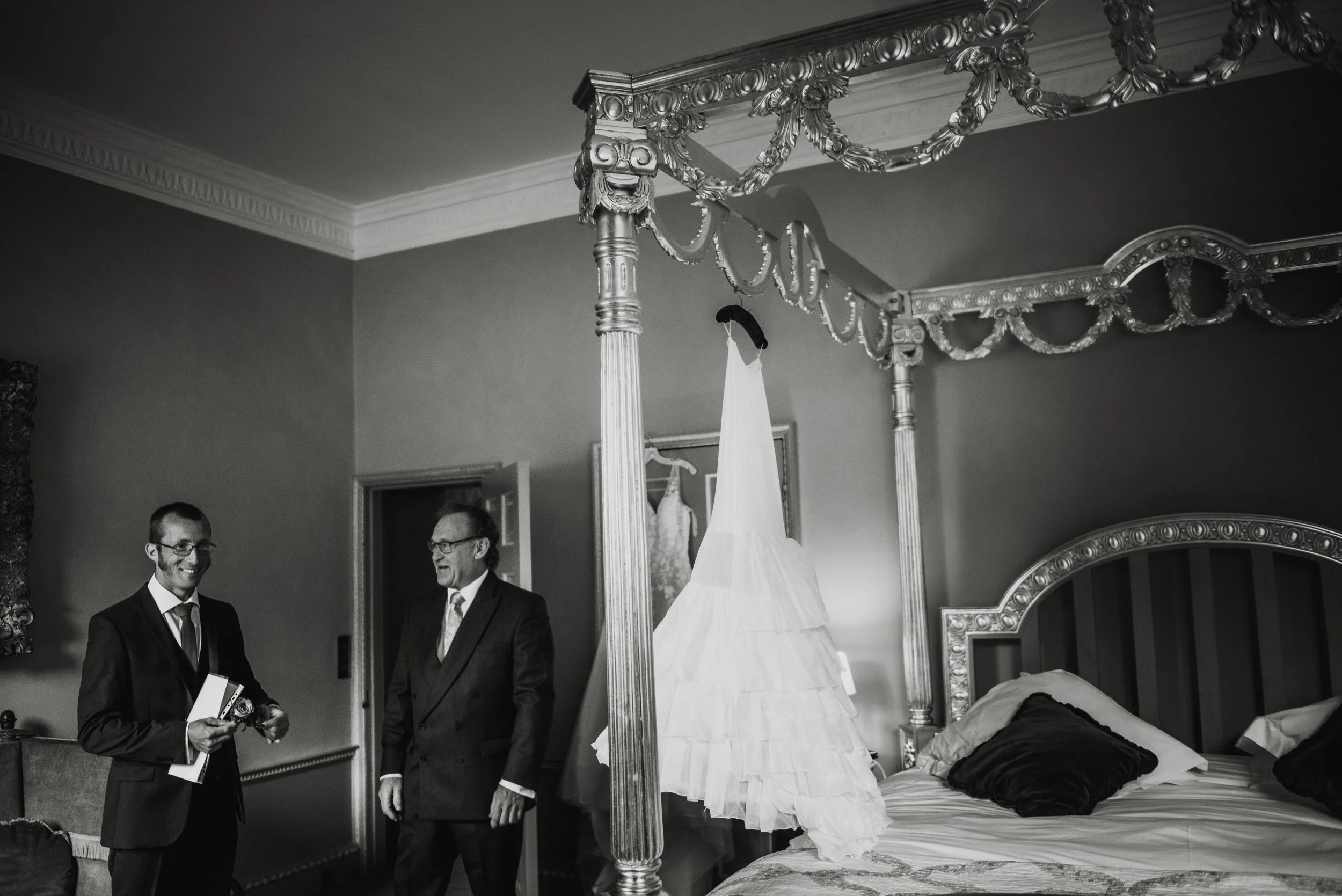 Two men in suits standing in a bedroom with a four-poster bed with a wedding dress hanging from one of the posts, in black and white.