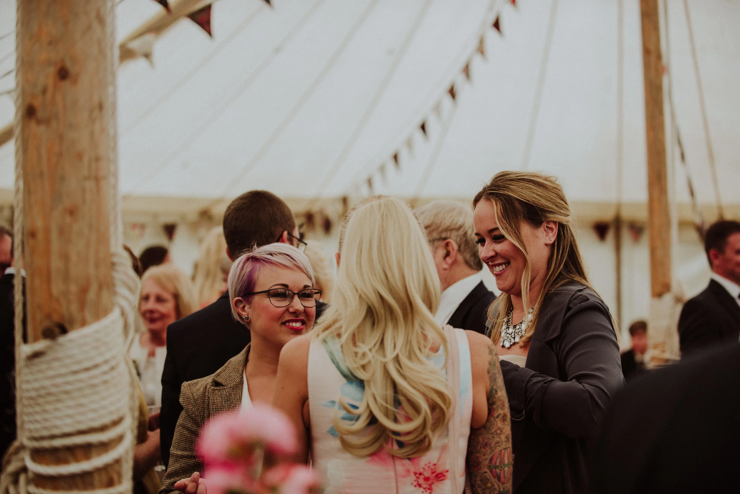 Group of smiling people talking at a celebration inside a large tent with wooden poles and bunting decorations.