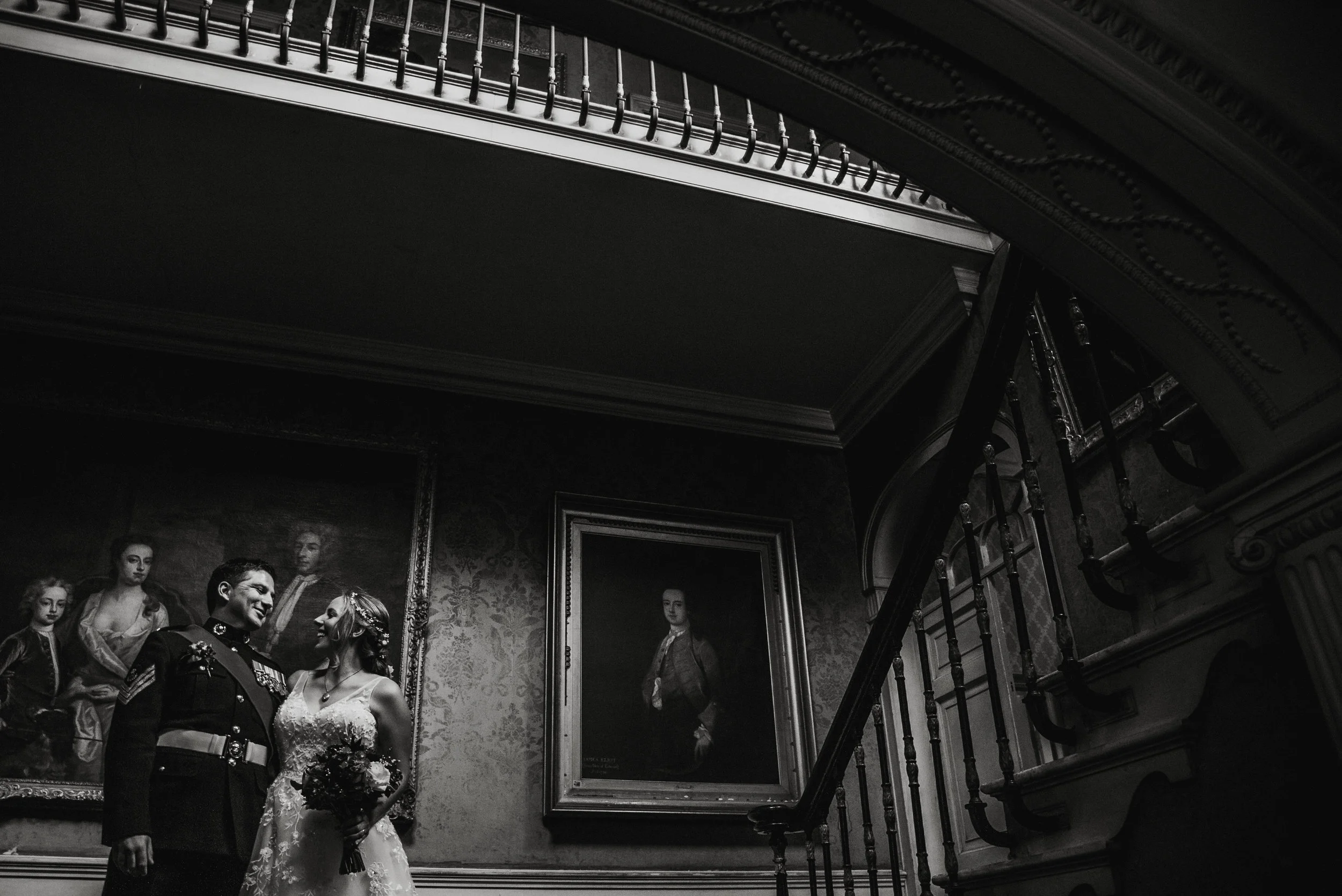 Black and white photo of a couple in wedding attire standing inside a historic building, with large framed classical portraits hanging on the wall behind them, a staircase on the right, and ornate decor.
