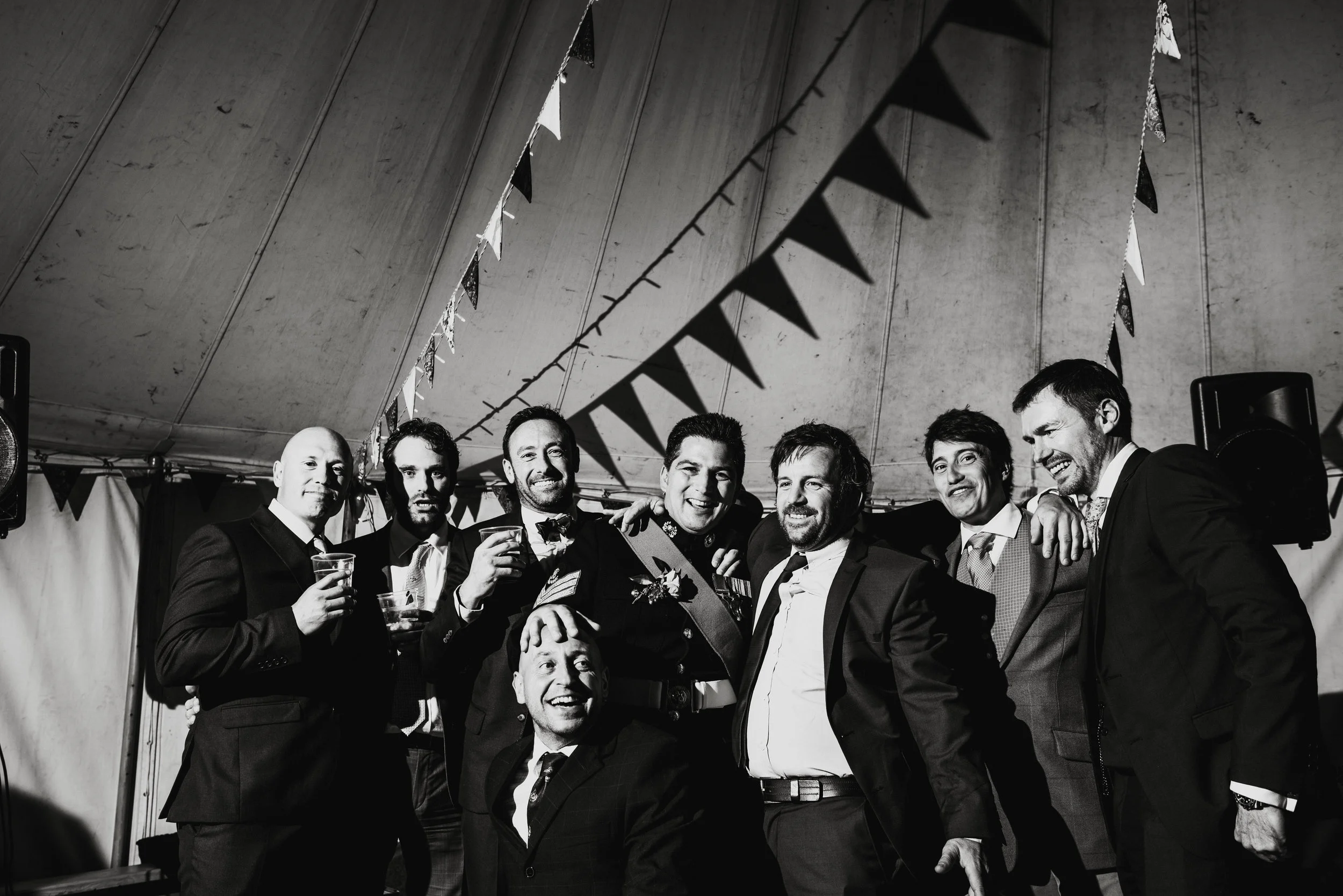 Group of men in formal suits celebrating at a party or wedding under a decorated tent.
