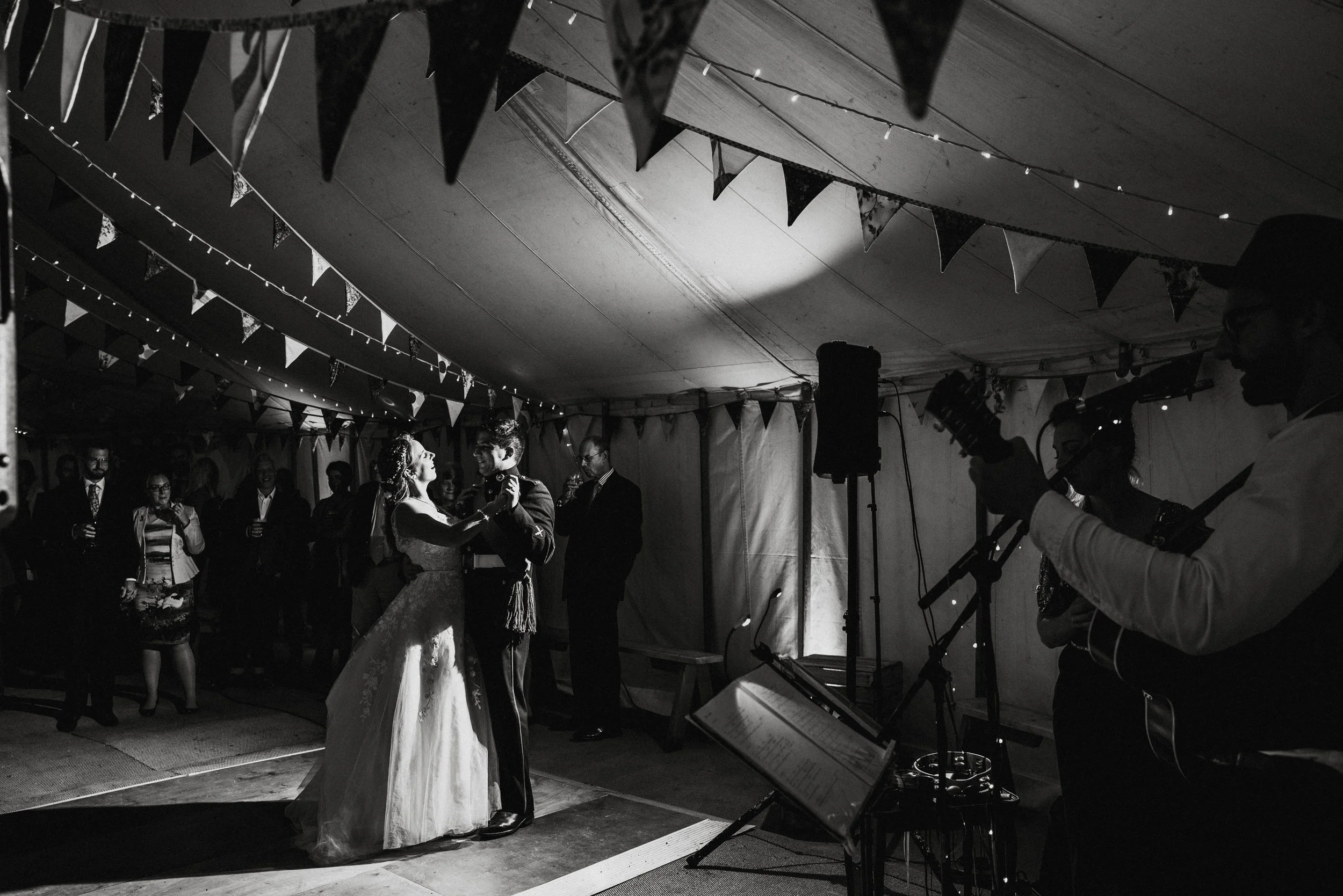 A black-and-white photo of a wedding reception, showing a bride and groom dancing under string lights and bunting. Guests are watching in the background, and musicians are playing in the foreground.