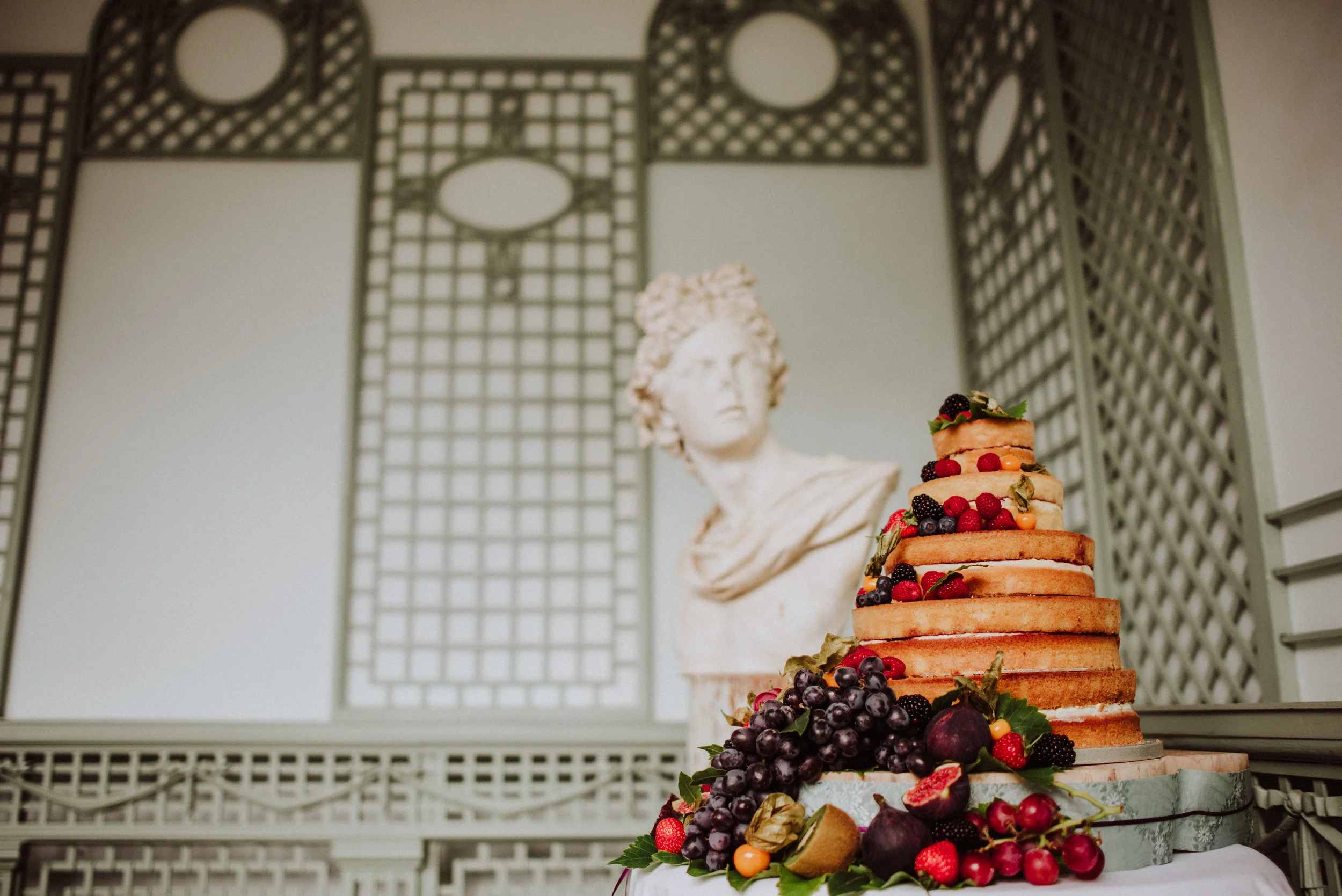 A multi-tiered naked cake decorated with fresh berries and grapes in front of a classical bust statue in a room with ornate lattice panels.