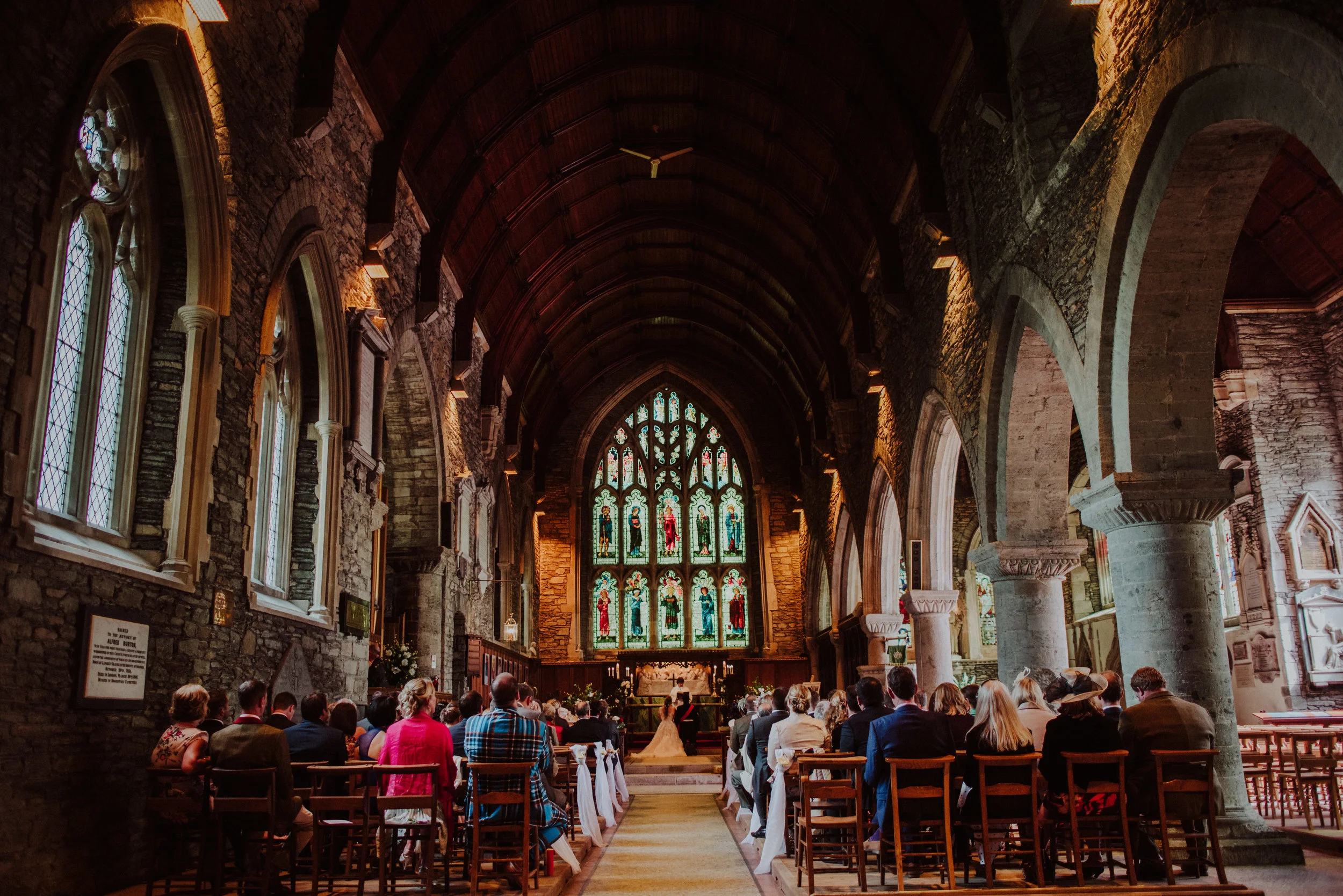 People attending a wedding ceremony inside a stone church with stained glass windows and wooden pews.