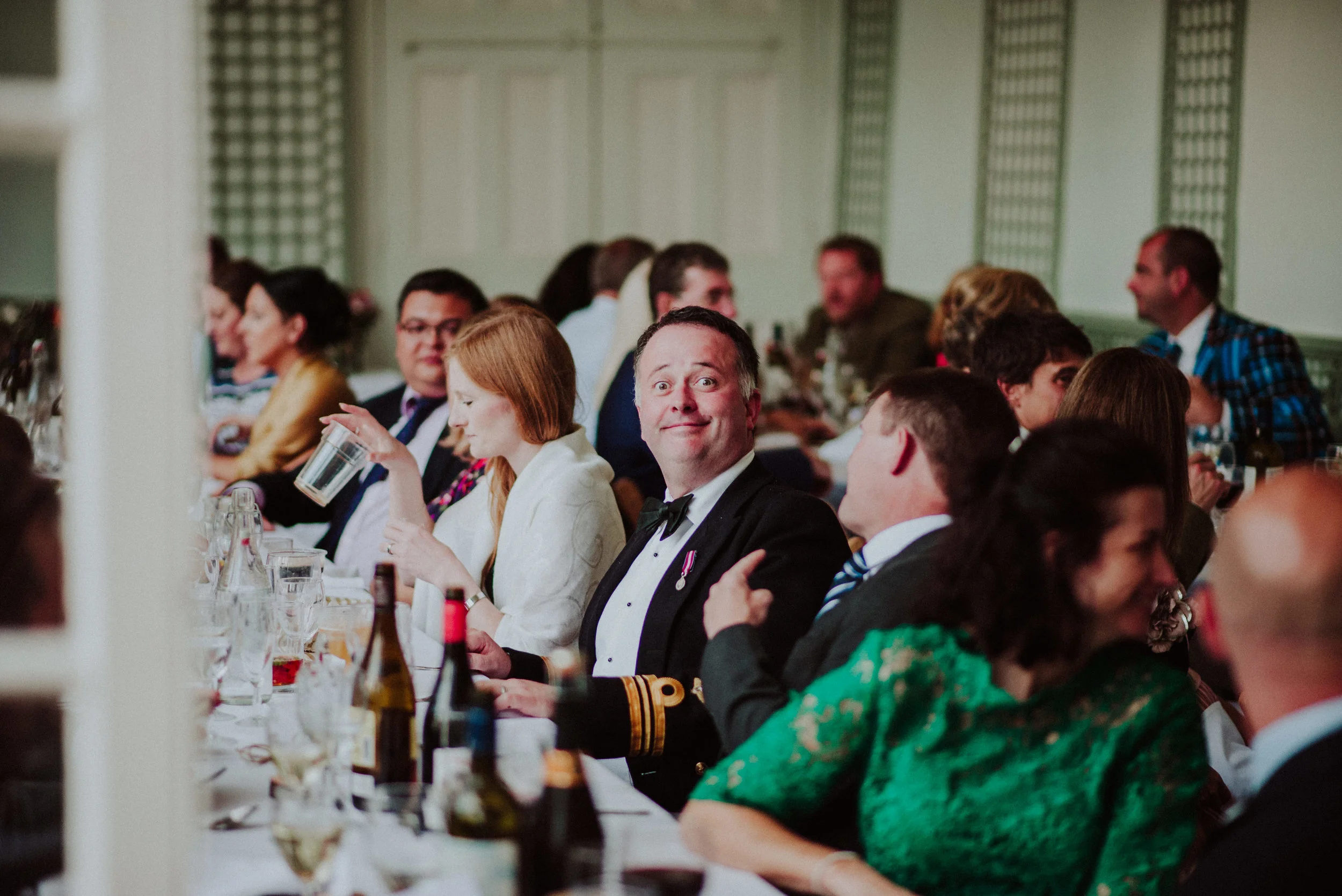 A group of people in formal attire sitting at a banquet table during a celebration or banquet event, with one man in a tuxedo smiling directly at the camera.