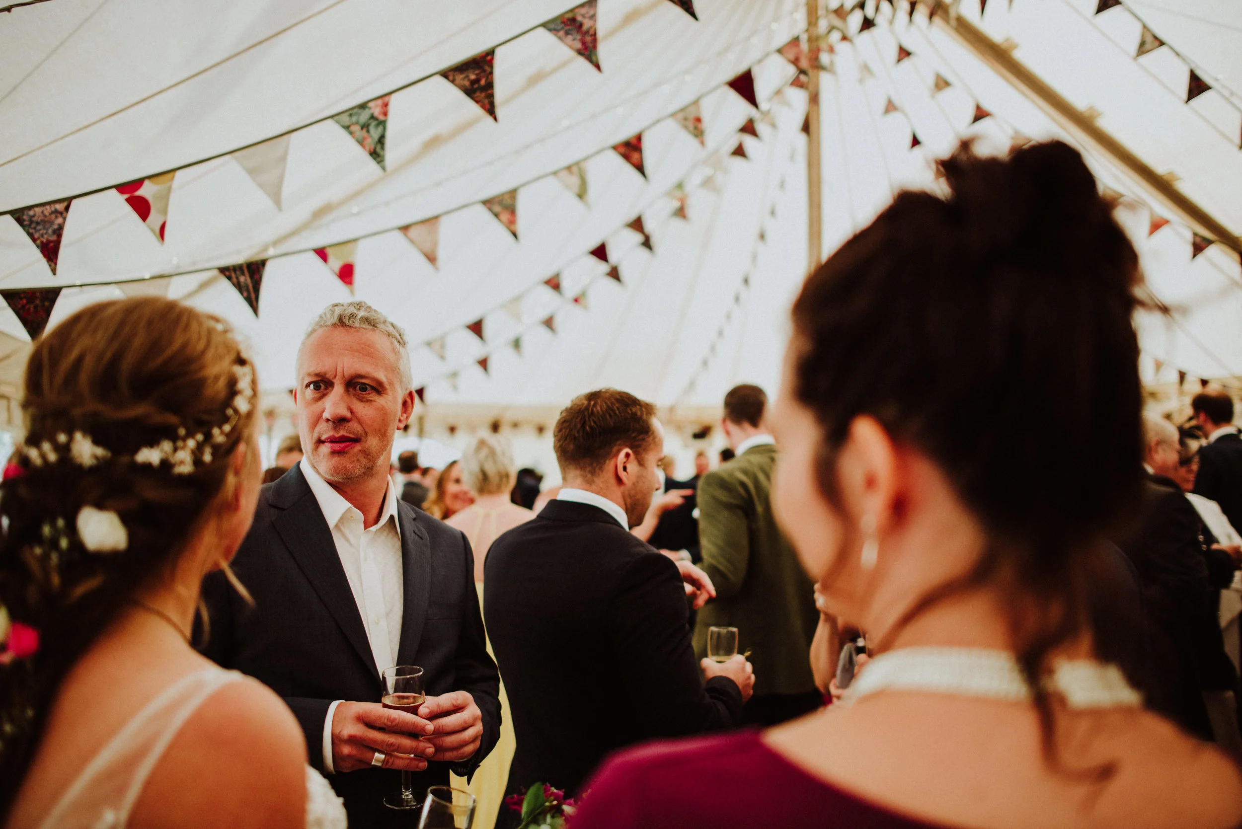 People at a wedding reception inside a decorated tent, engaged in conversation and holding glasses of champagne.