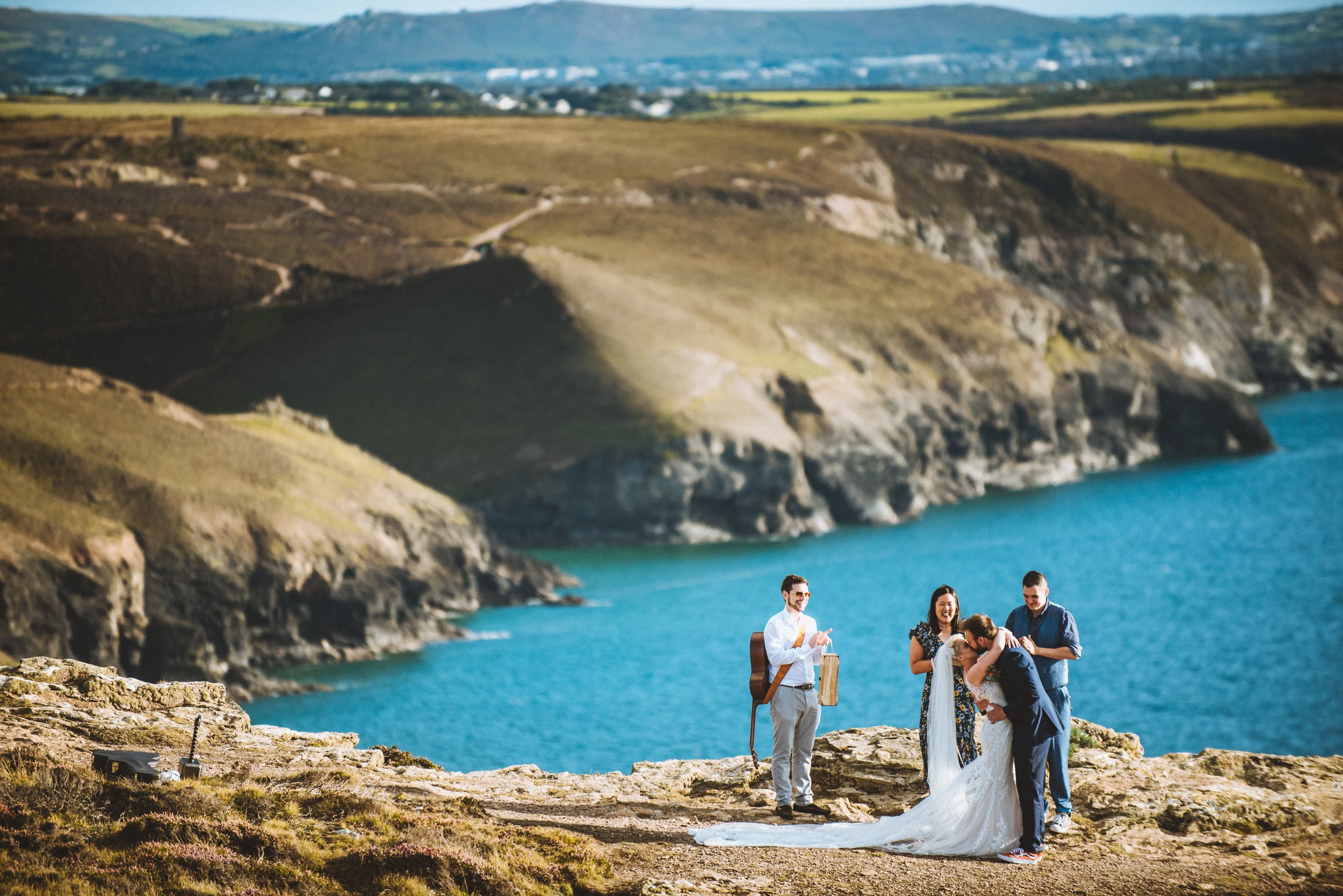 A couple getting married outdoors near a body of water with rolling hills in the background, surrounded by two friends or family members.