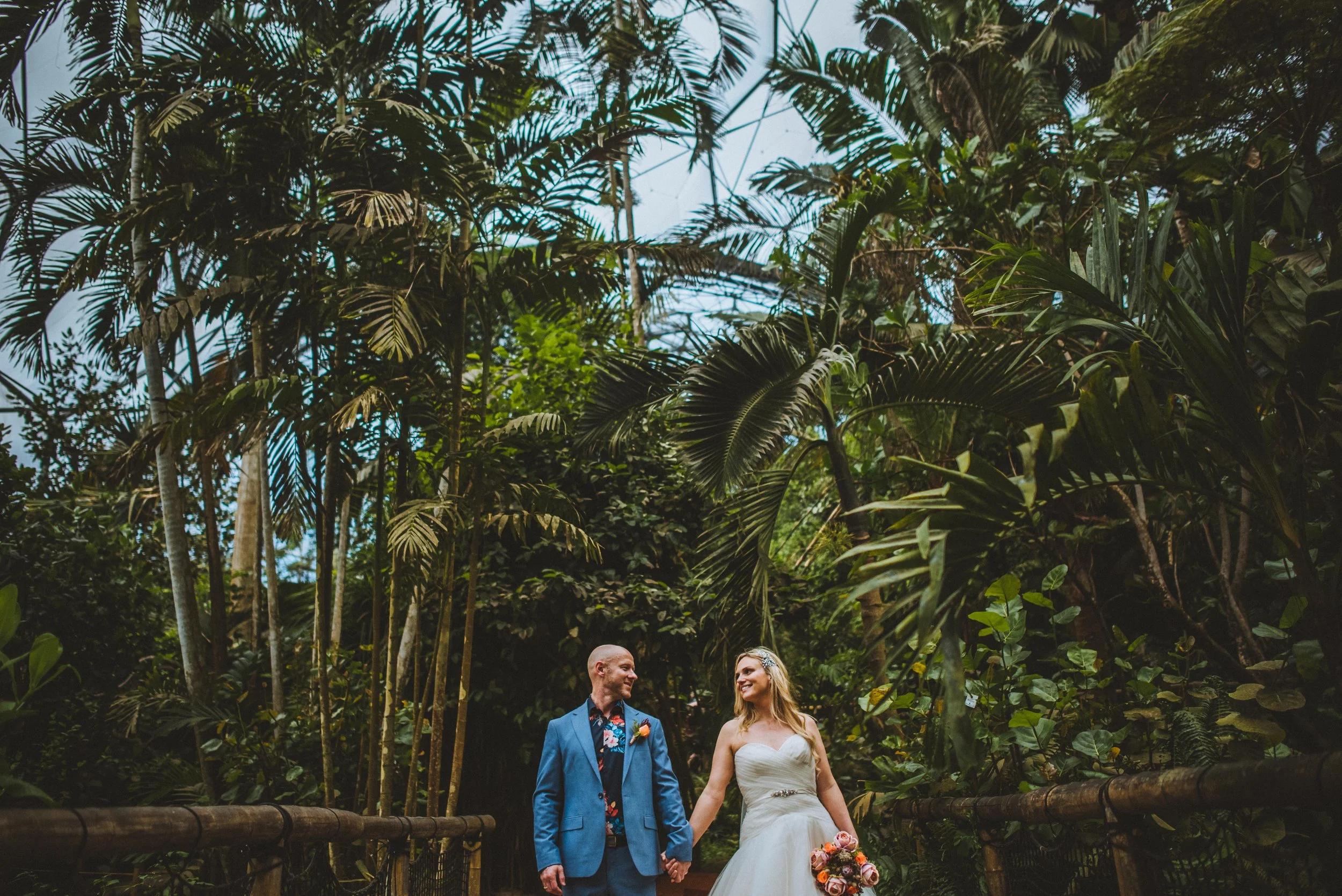 A bride and groom walking hand in hand through a lush tropical forest, with the bride holding a bouquet of flowers and both smiling at each other.