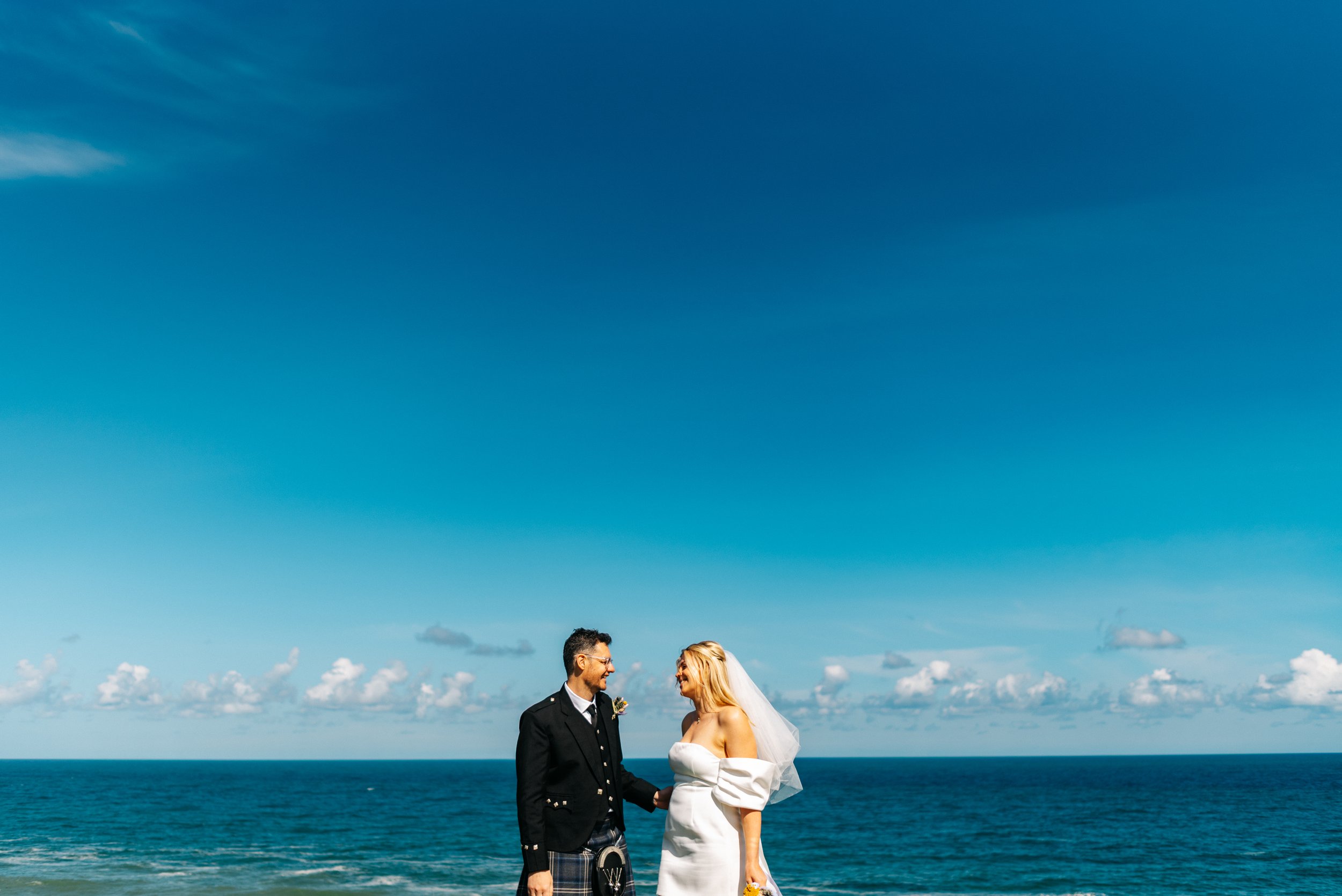 A bride and groom holding hands on a beach with the ocean and blue sky in the background.