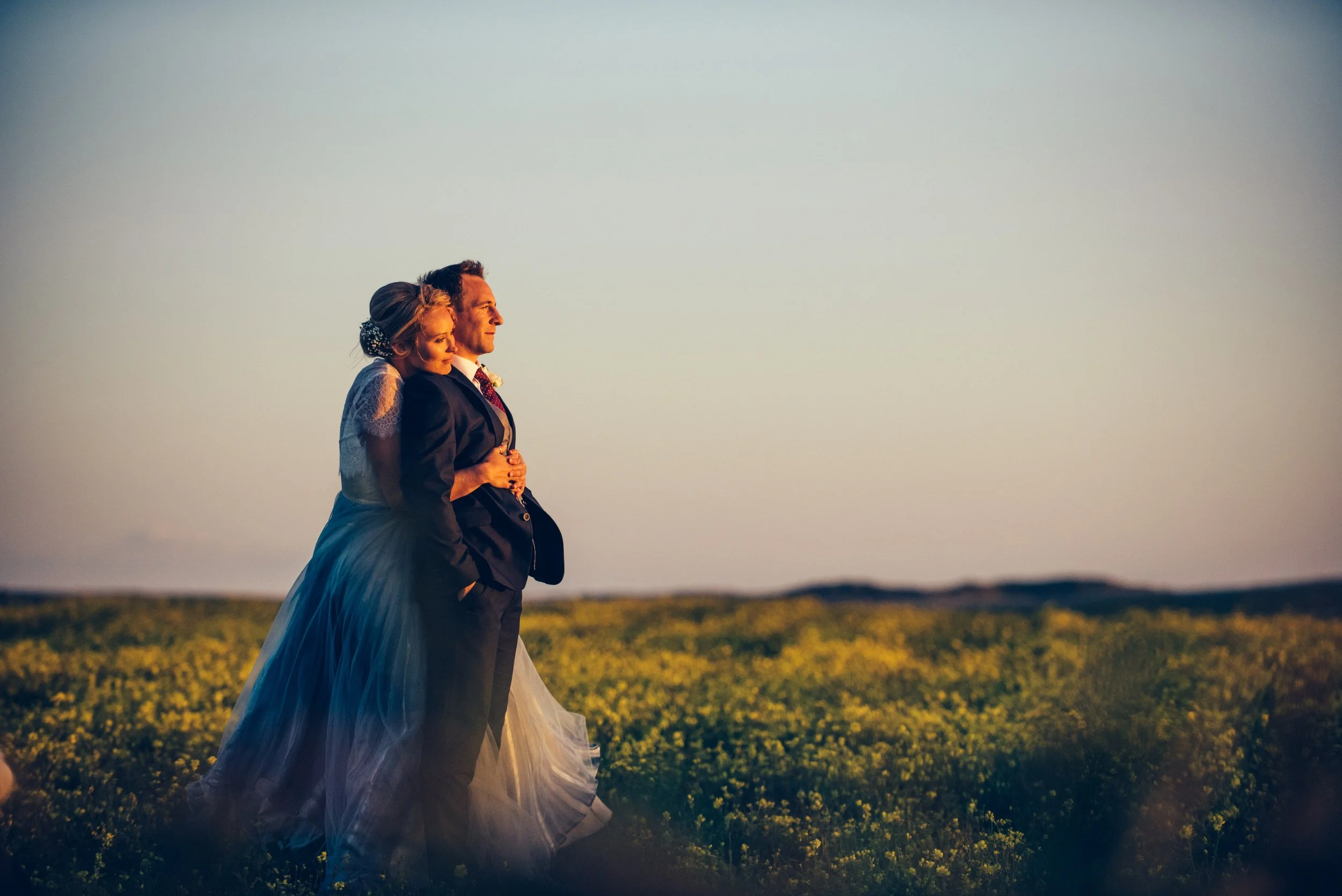 A bride and groom standing in a field during sunset, with the bride leaning on the groom's shoulder and holding his arm, wearing wedding attire.