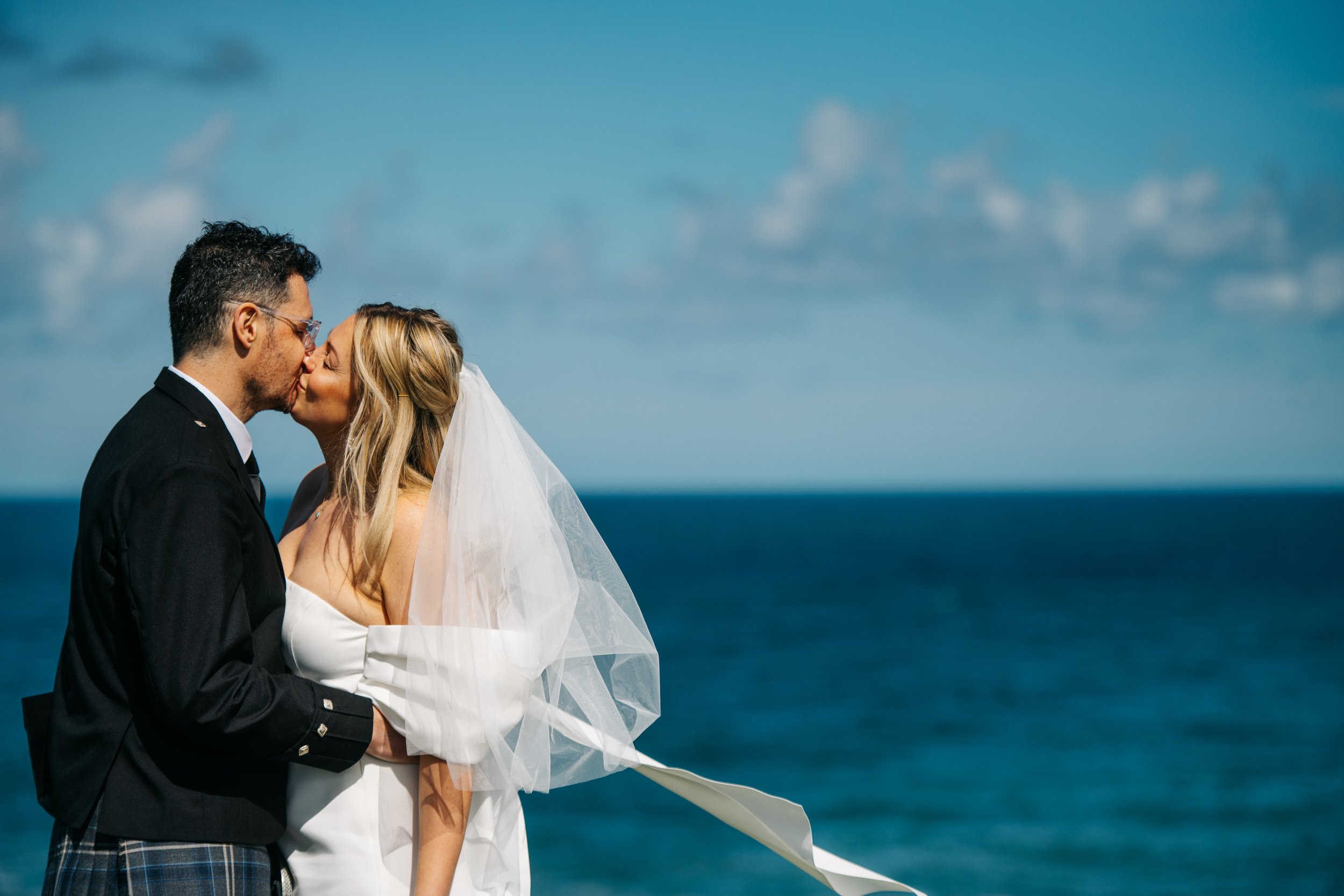 A bride and groom share a kiss by the ocean on their wedding day, with a blue sky and clouds in the background.