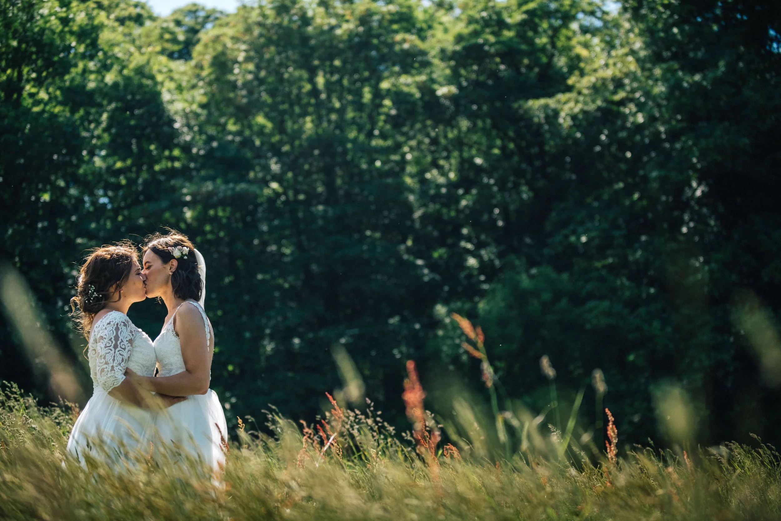 Two brides in wedding dresses sharing a kiss in a grassy field with a dense, green forest in the background.
