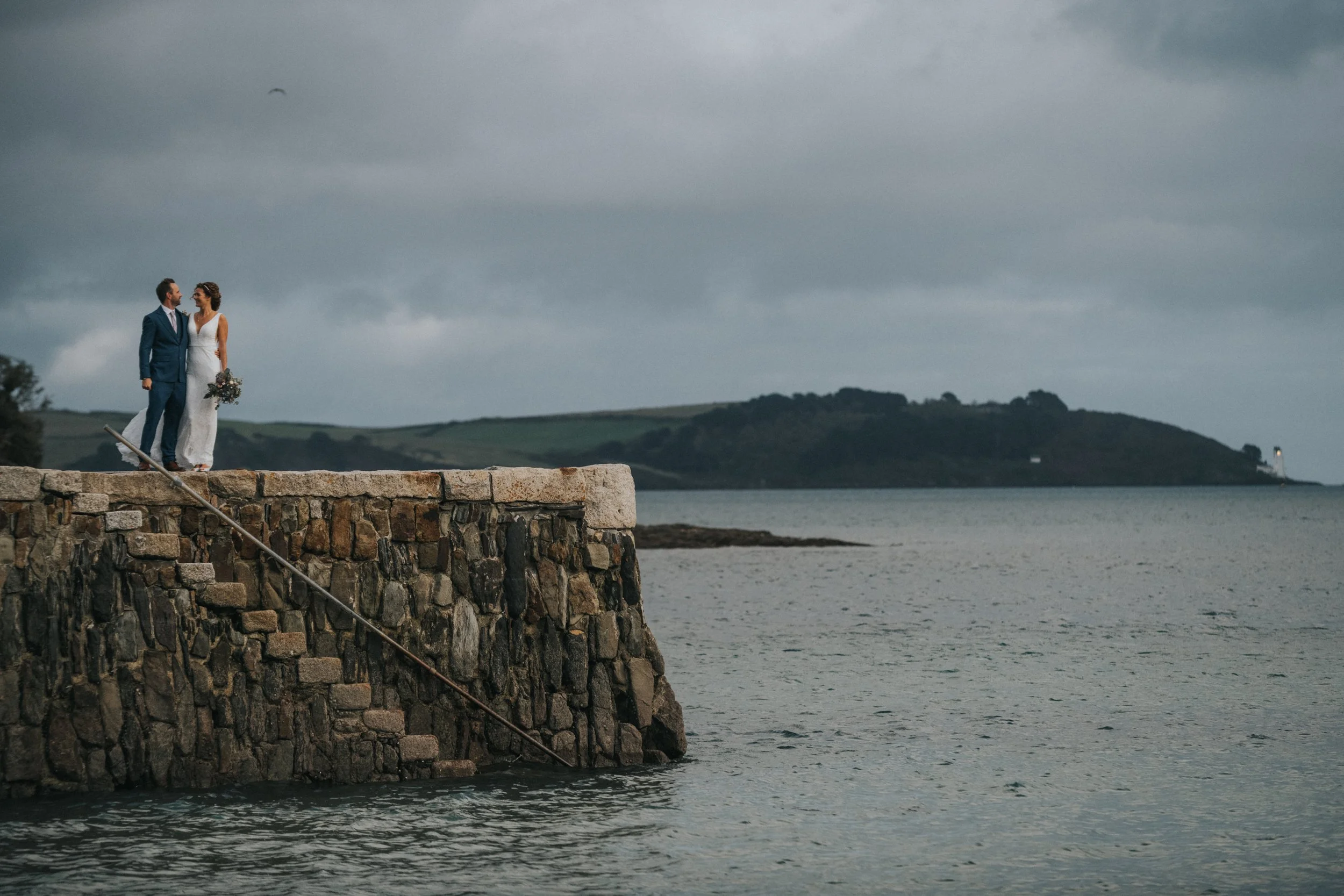 A bride and groom standing on a stone pier by the water on a cloudy day, with a lighthouse visible in the distance on an island.