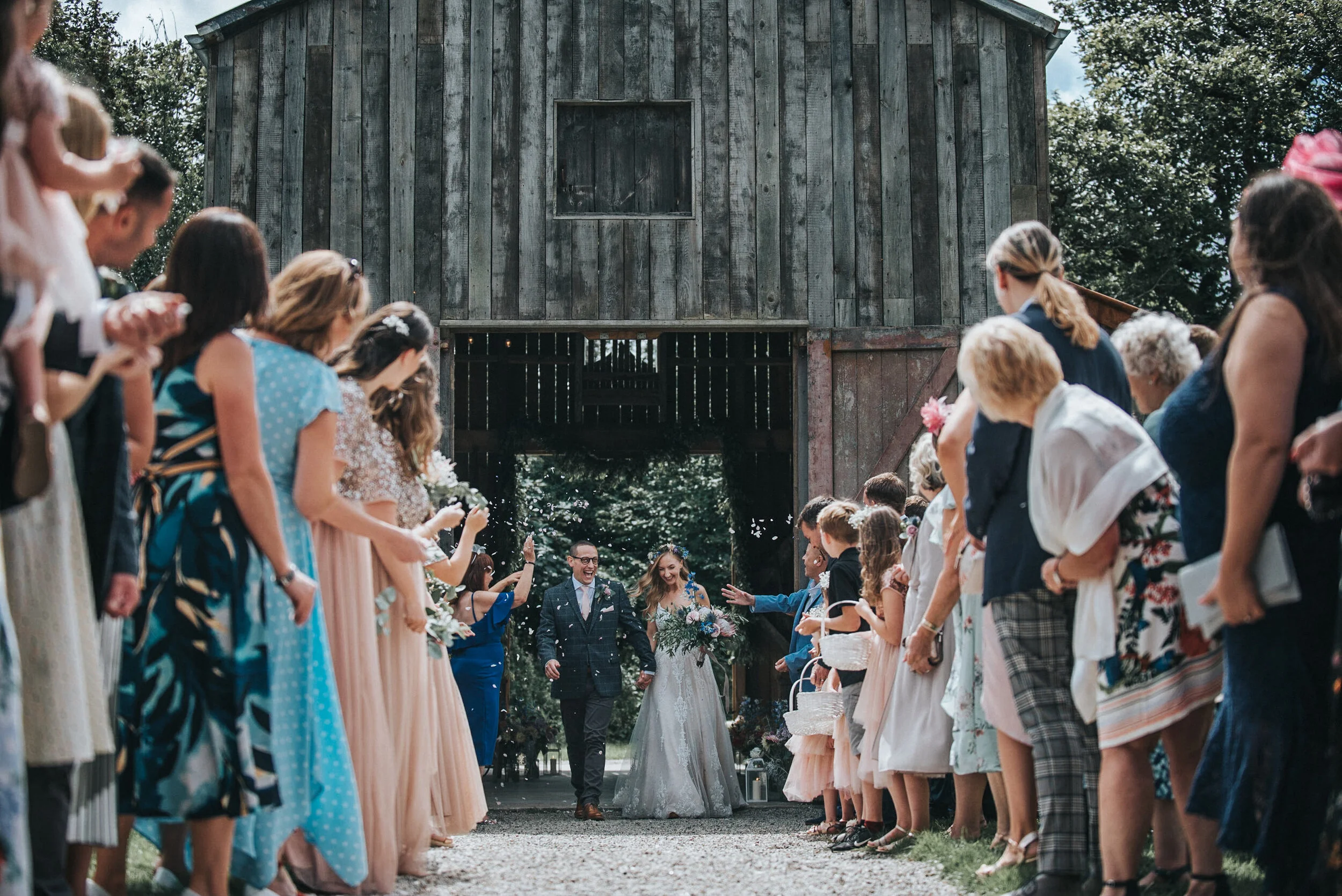 Nancarrow Farm Wedding Photography - A wedding ceremony exit with a bride and groom walking away from a rustic barn, surrounded by guests celebrating outdoors.