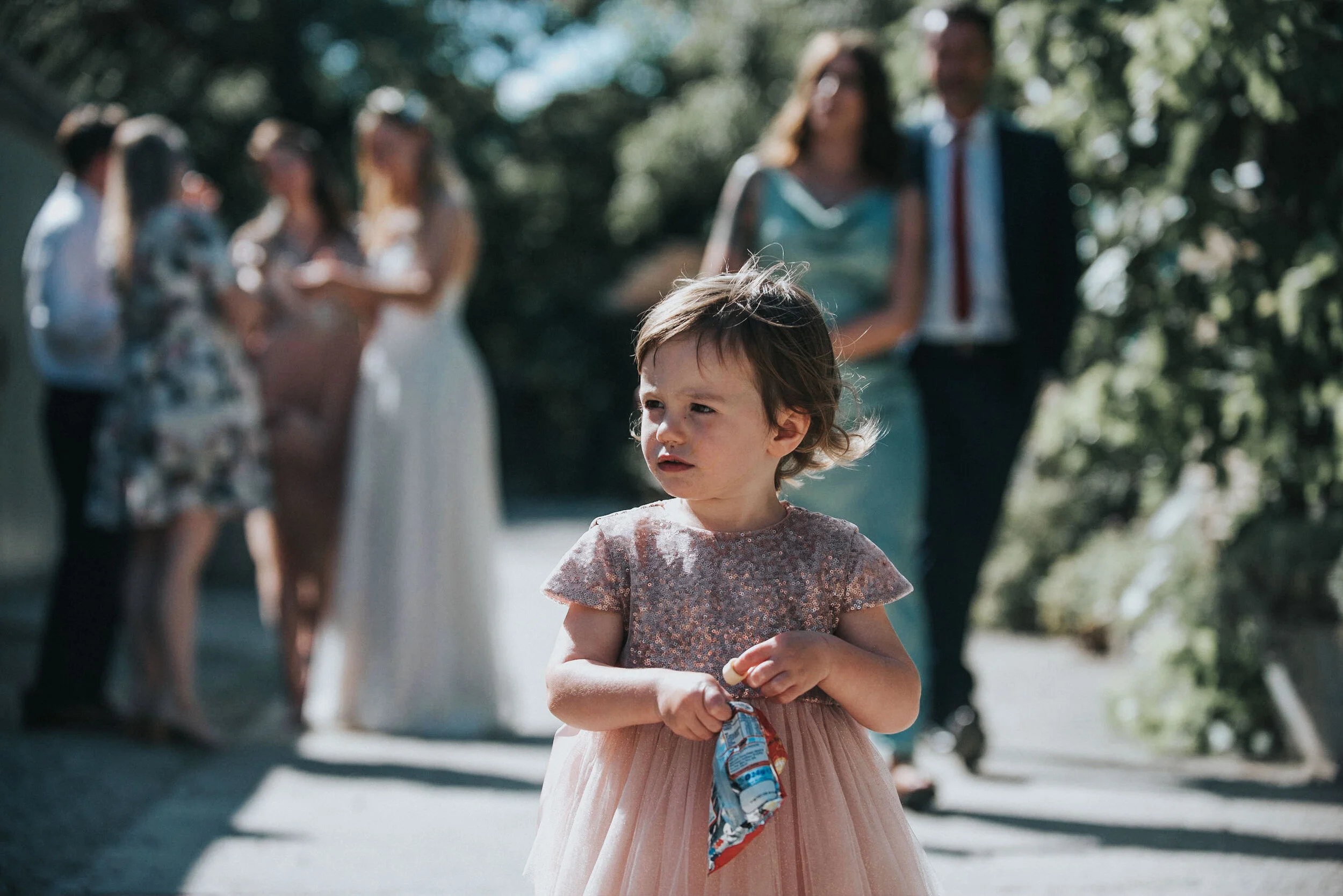 A young girl in a pink dress holding a snack stands outside while a group of wedding guests in formal attire, including a bride, groom, and others, are blurred in the background.