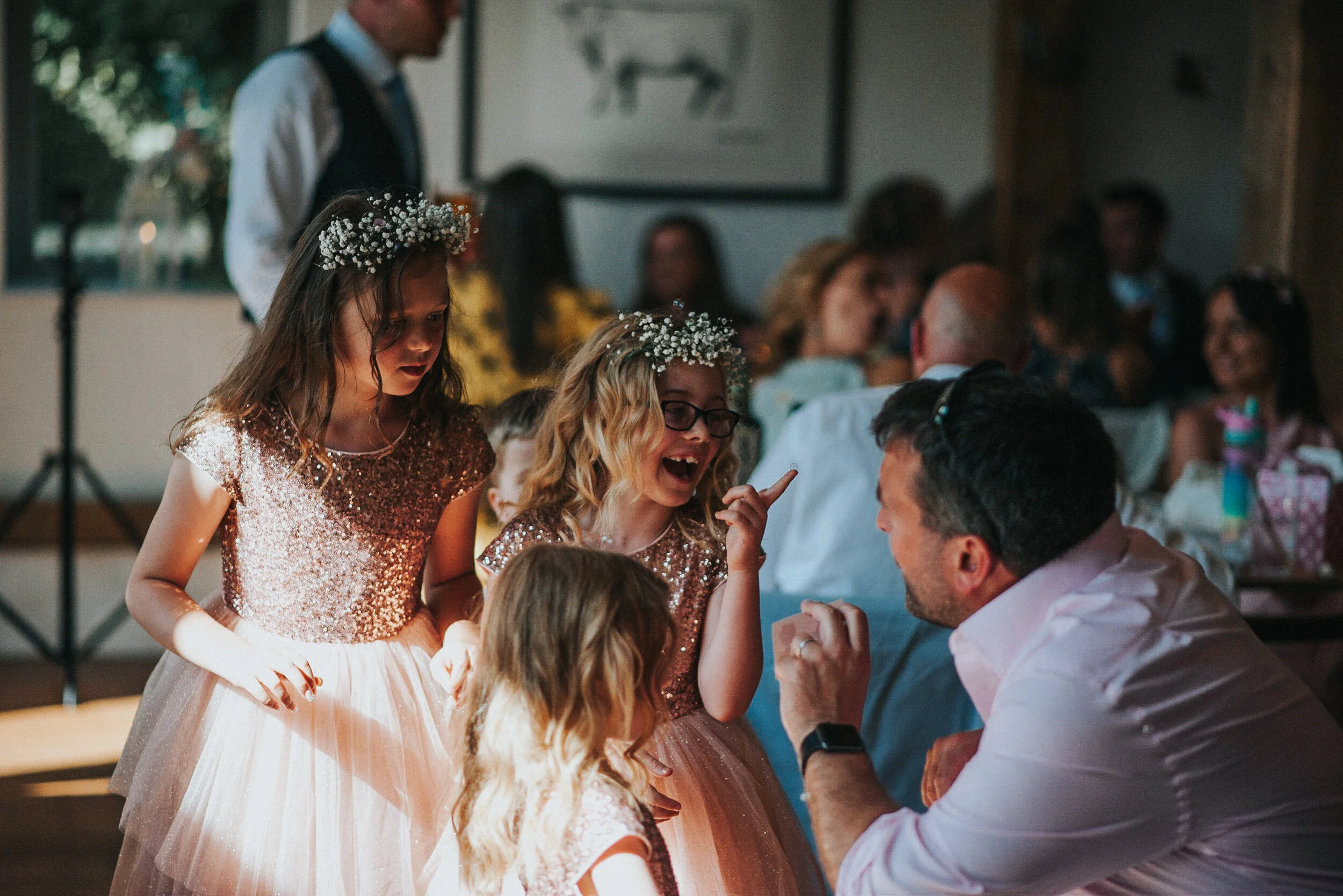 Children and adults at a wedding reception, girls wearing floral crowns and dresses with sequined tops, engaging in conversation with a man who is smiling and talking to them.
