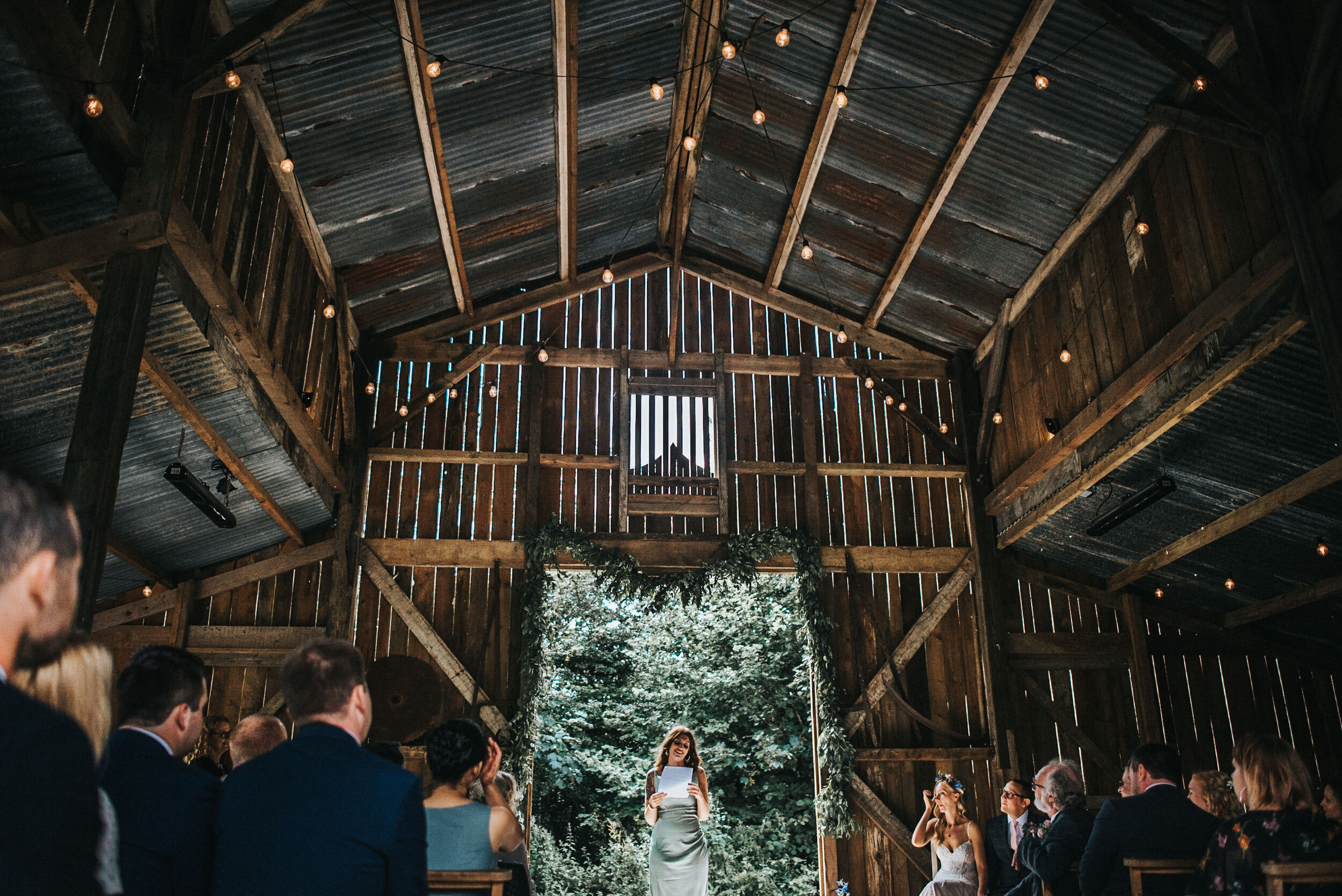 A wedding ceremony inside a rustic barn with wooden walls and ceiling, string lights hanging overhead, and a woman reading vows at the altar decorated with greenery, while guests are seated on either side.