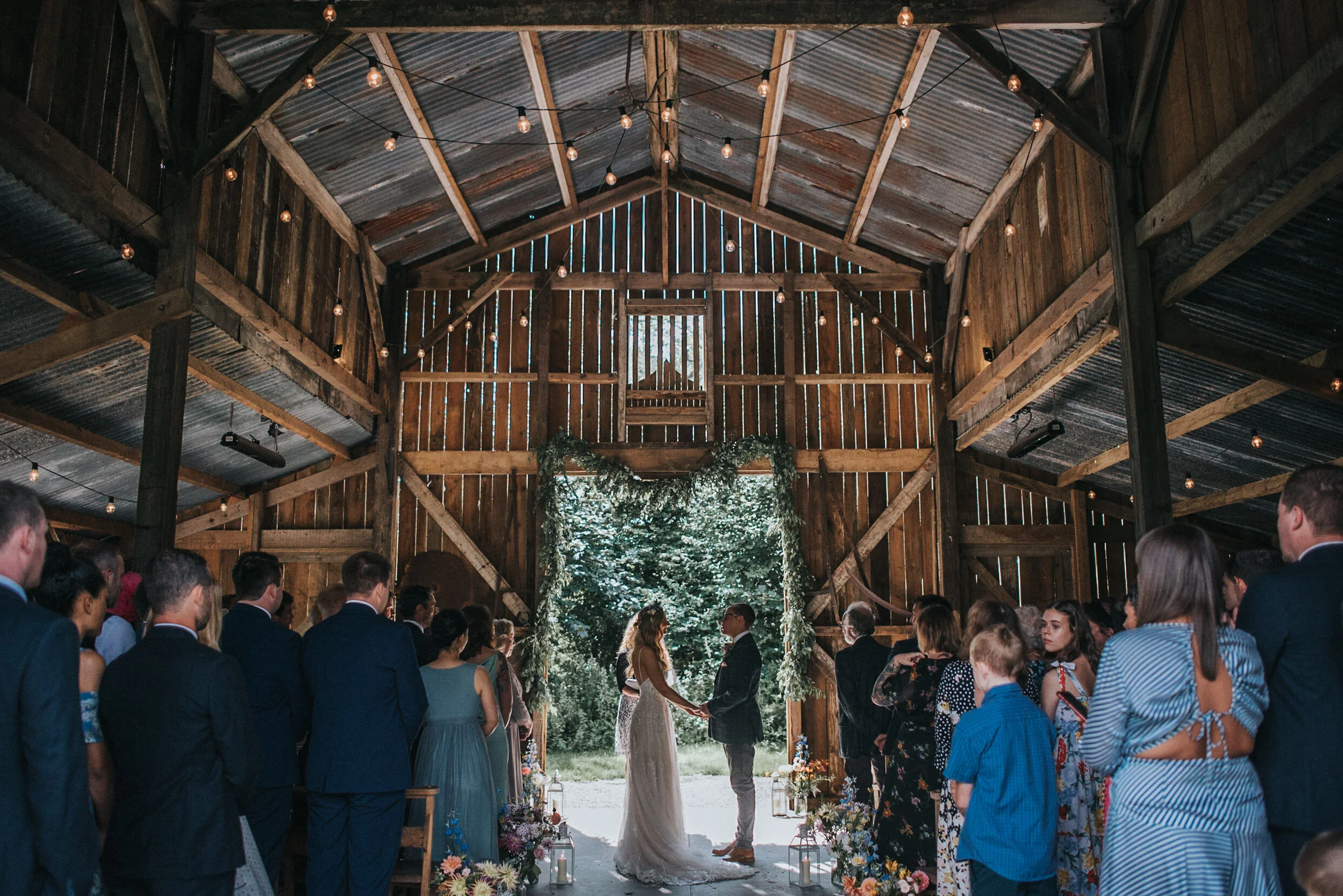 A wedding ceremony taking place inside a rustic barn with wooden walls and ceiling, decorated with string lights and greenery arch, with the bride and groom exchanging vows in front of an outdoor forest backdrop.