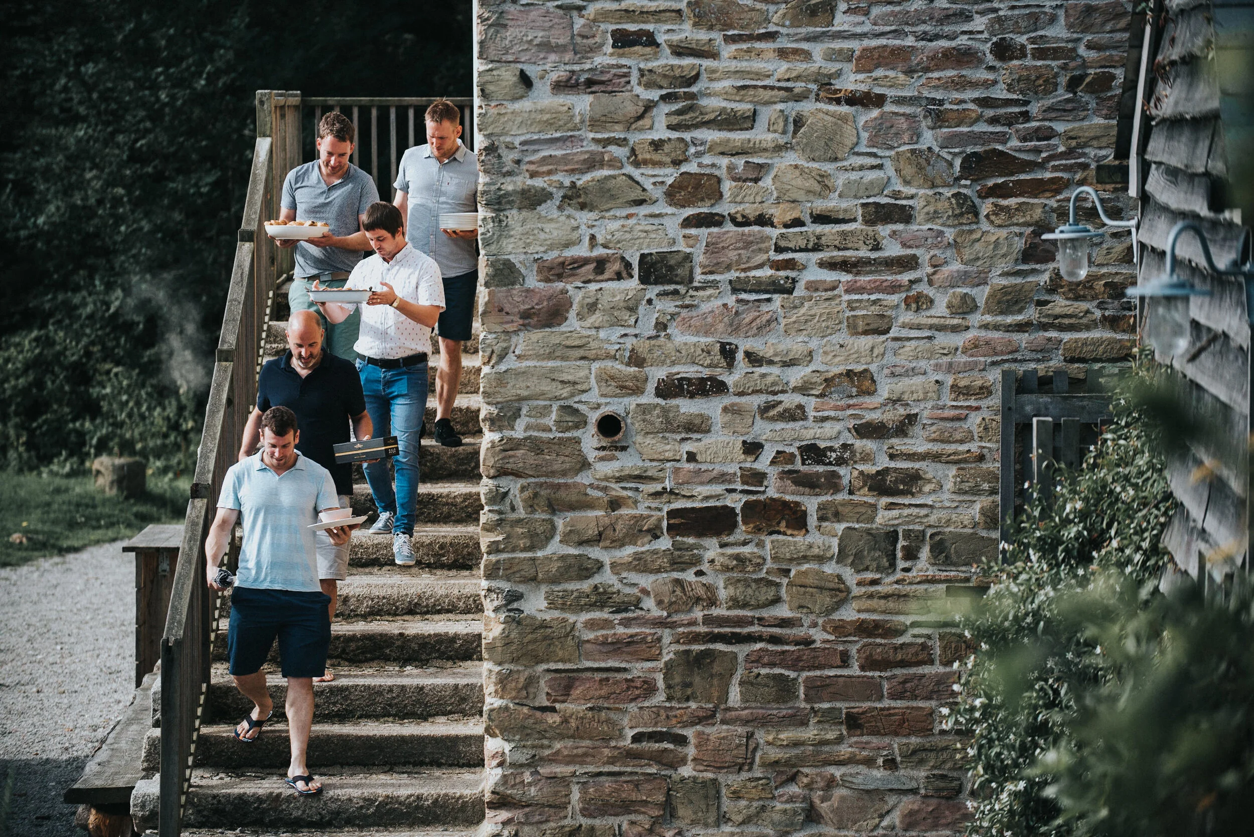 A group of six men descending outdoor stone stairs while holding plates of food, with greenery and a brick wall in the background.