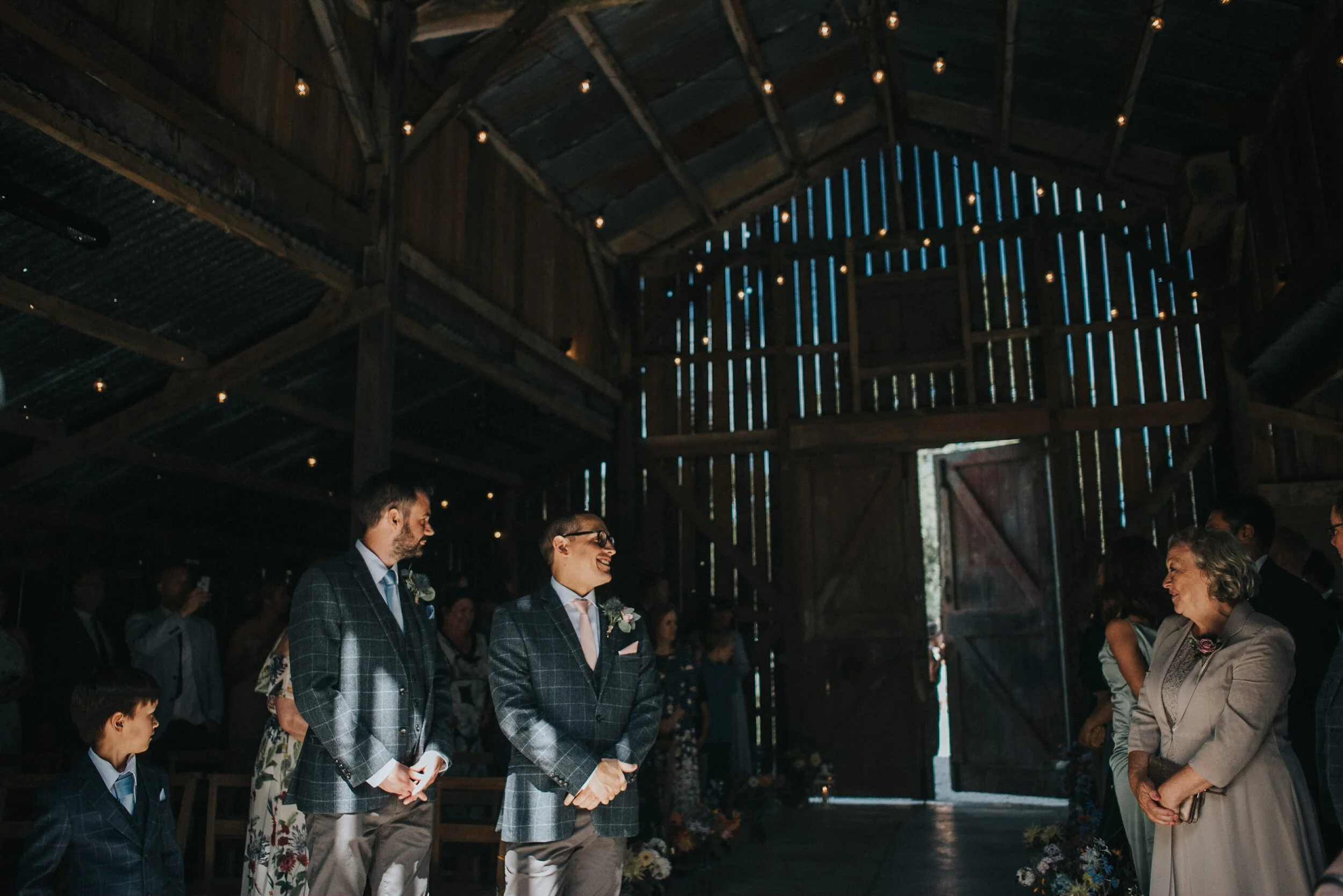 A wedding ceremony inside a rustic barn with natural light streaming through wooden gaps. The bride and groom stand with guests, who are dressed formally, as the officiant prepares to conduct the ceremony.