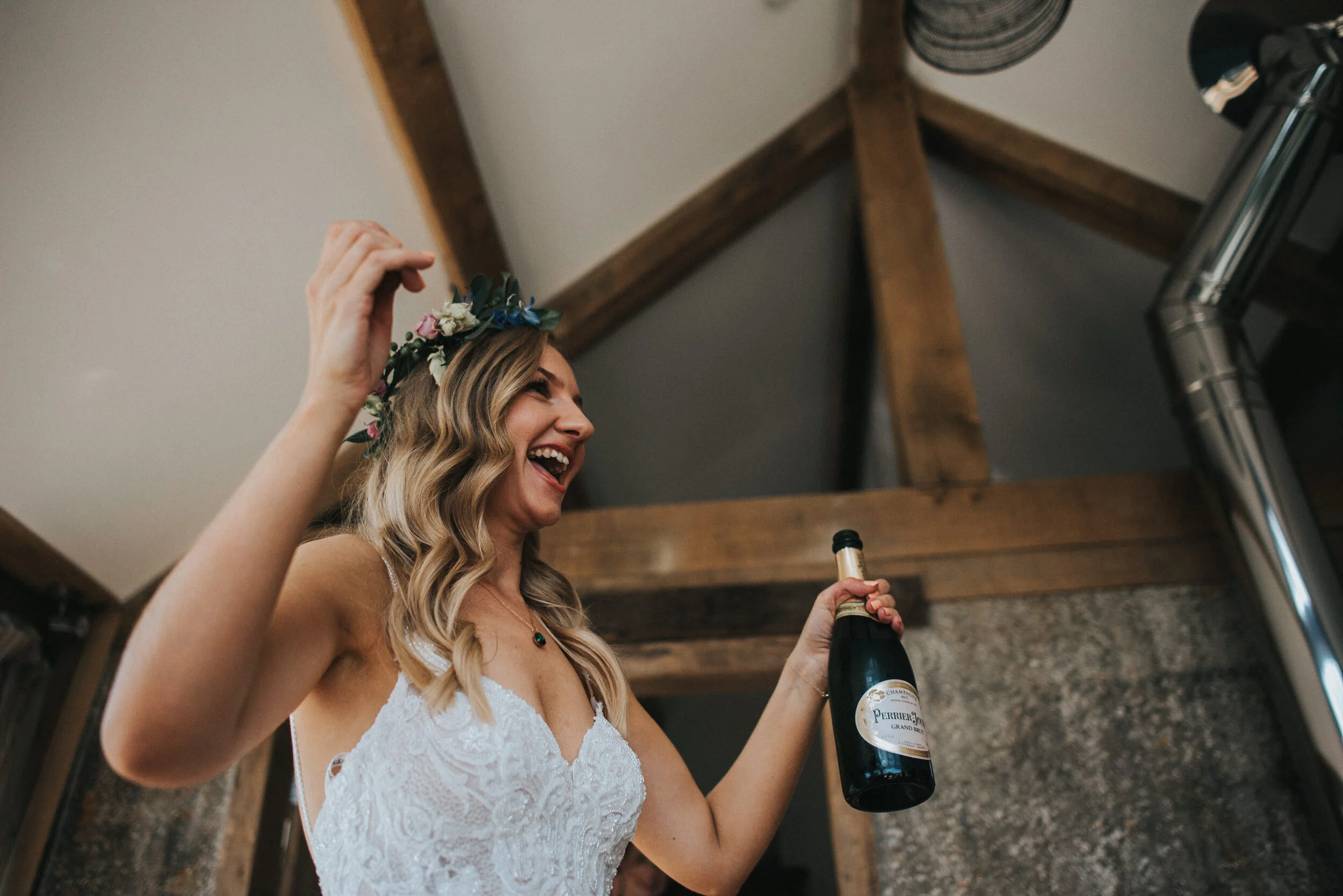 A woman in a wedding dress and a floral crown is celebrating with a bottle of champagne.