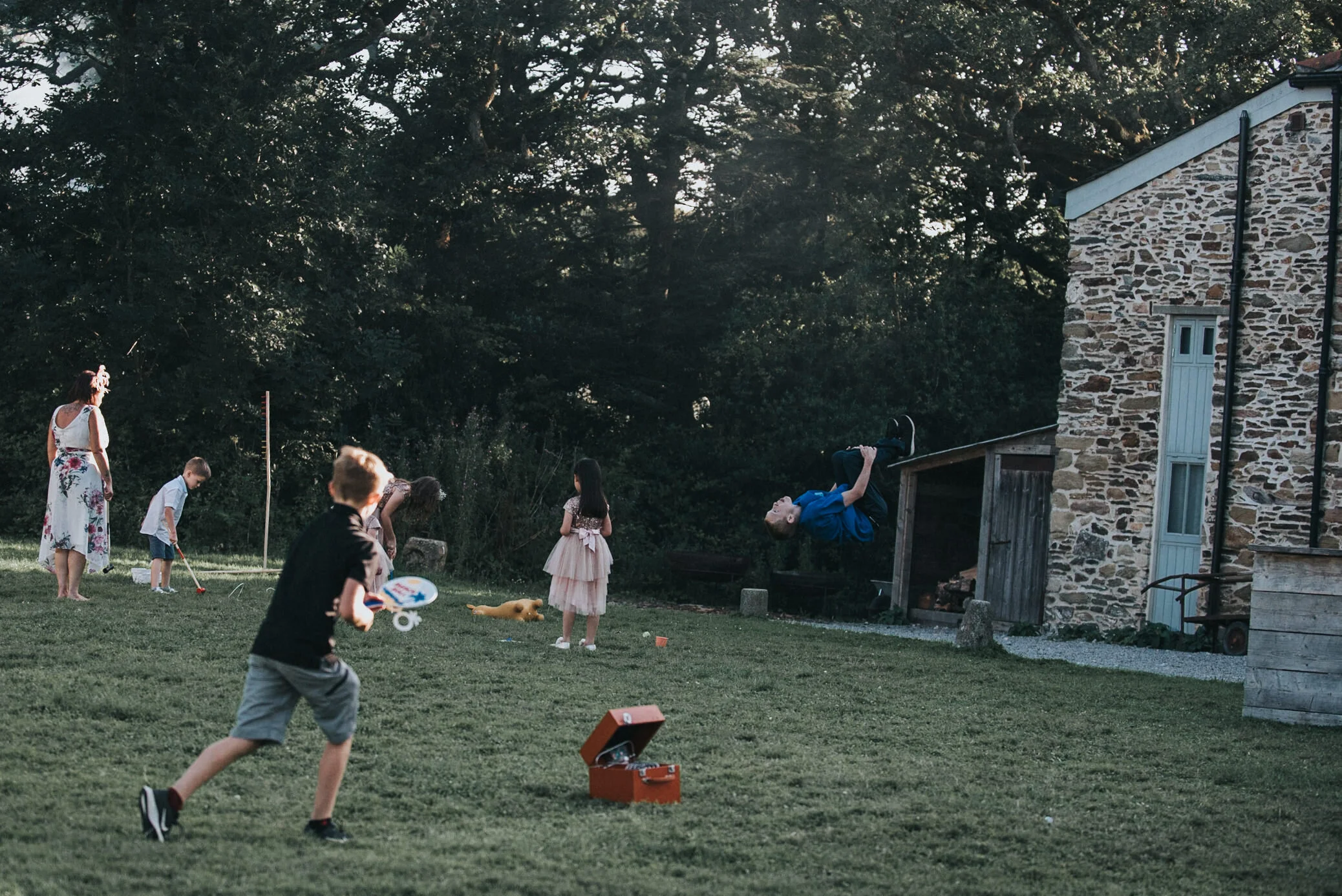 Children and adults playing and engaging in outdoor activities on a grassy yard near a stone building, with a person flipping in mid-air on a trampoline, while others are playing with a toy and walking around.
