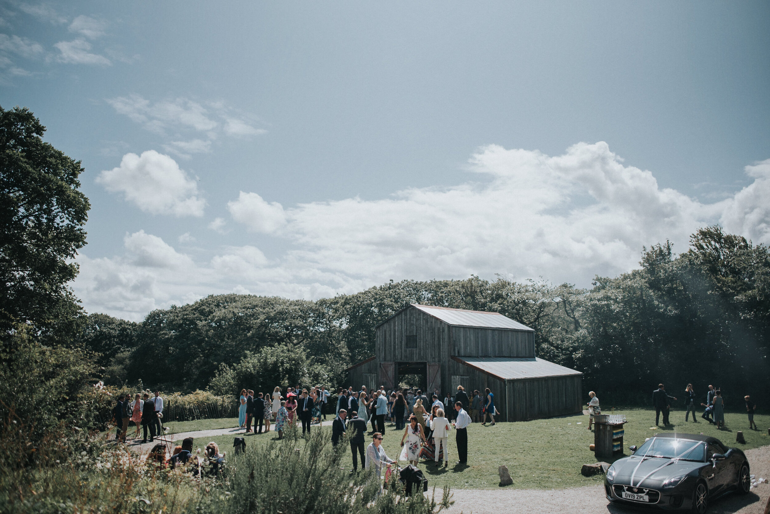 Outdoor gathering of people near a rustic wooden barn on a sunny day with scattered clouds, surrounded by trees and greenery.