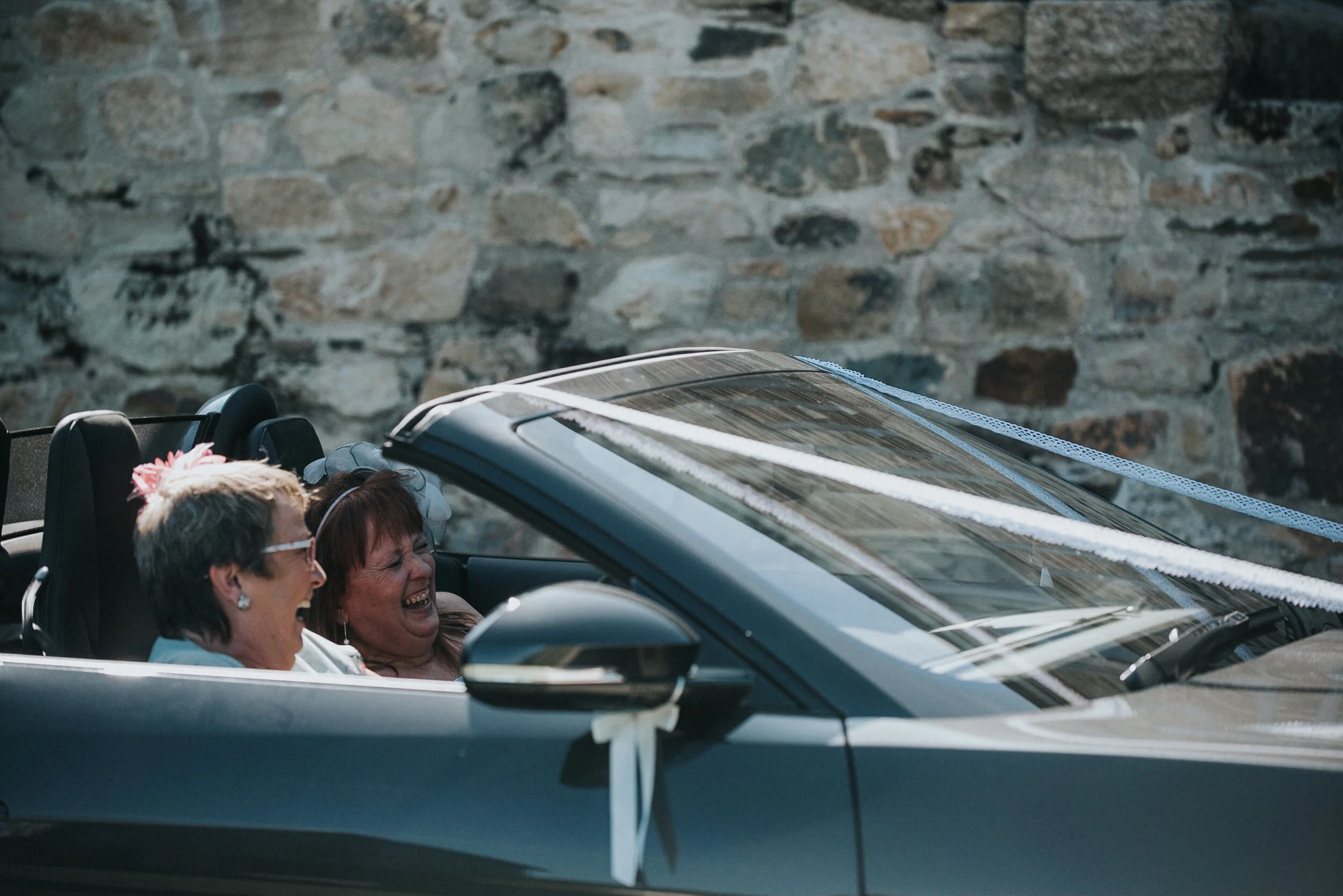 Two women laughing and smiling while riding in a convertible car decorated with white ribbons and flowers, in front of a stone wall.