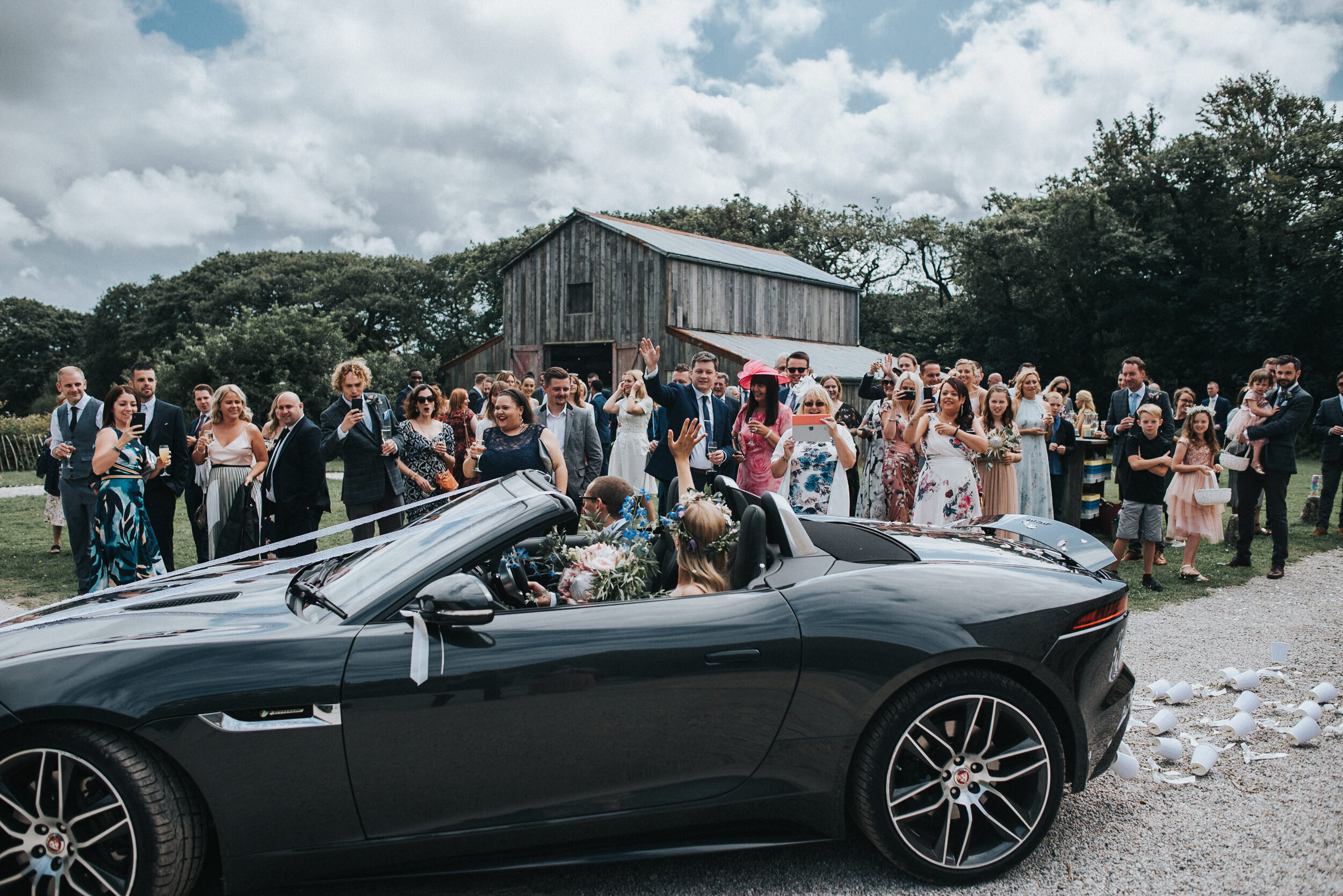 A wedding celebration at Nancarrow Farm in Cornwall with guests standing around a black convertible car that is decorated. 