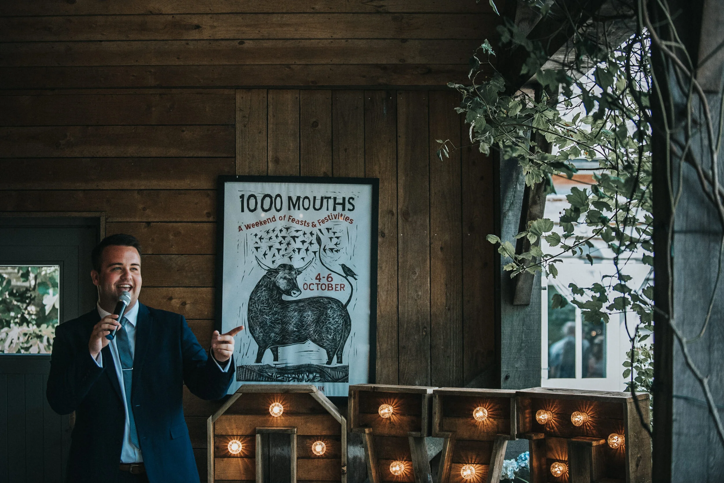 A man in a dark suit holding a microphone and speaking with a smile, standing inside a wooden building, with a poster behind him advertising an event called "1000 MOUTHS" featuring a bison illustration and scheduled for October 4-6.