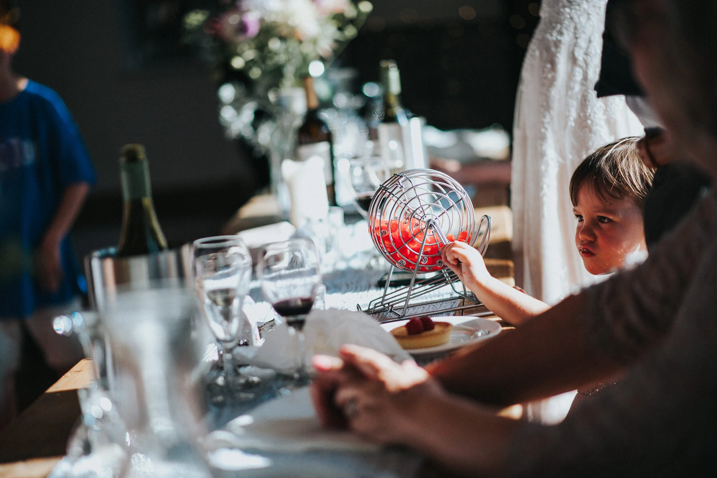 A young boy reaching into a bingo cage filled with red balls at a celebration or party, with a table set with wine glasses, bottles, and a small dessert on a plate nearby.