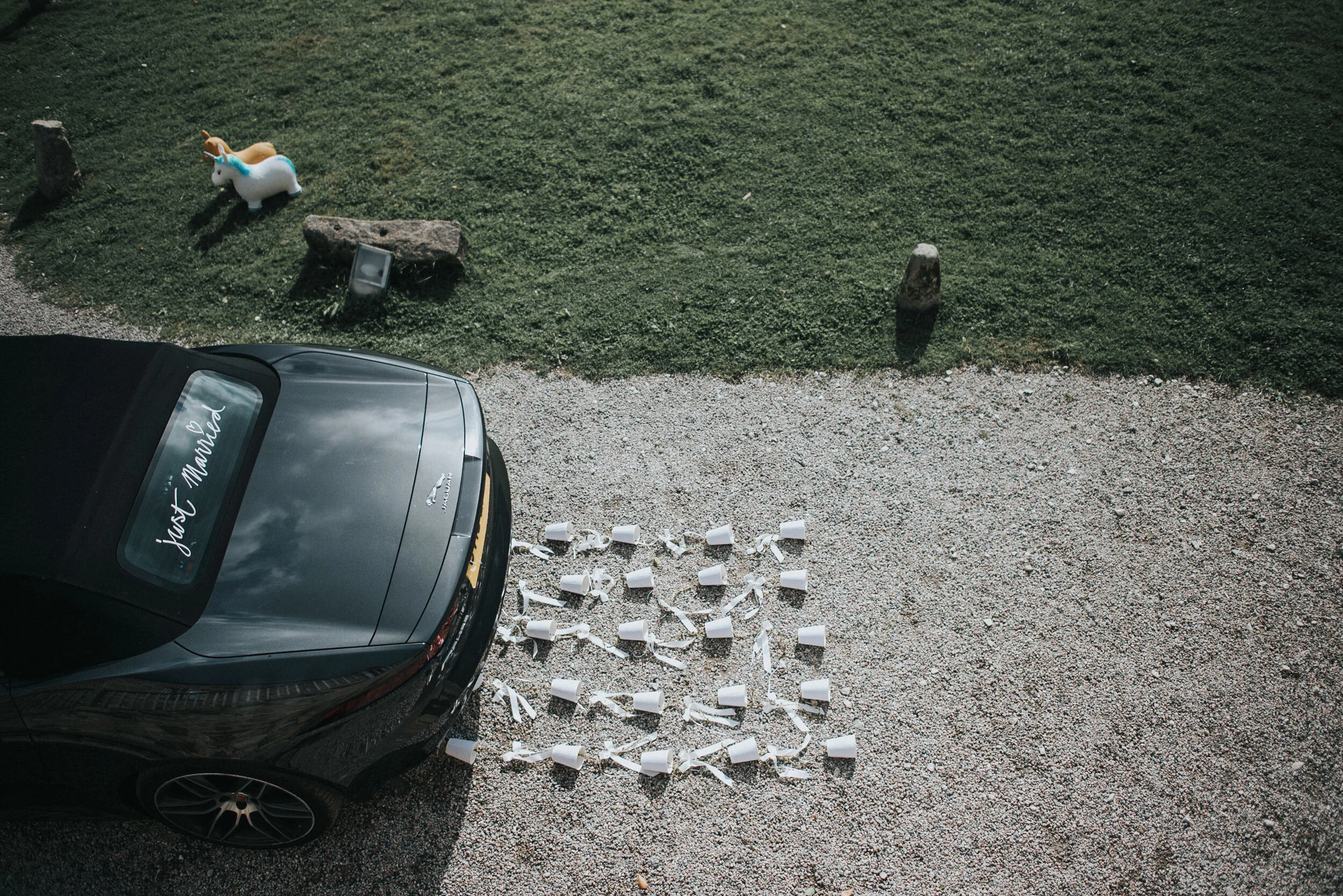 Overhead view of a black car parked on gravel with 25 white coffee mugs arranged on the ground to form a heart shape next to the car. There is a sign on the car's rear window that reads 'Just Married'. In the background, there is a patch of grass wit